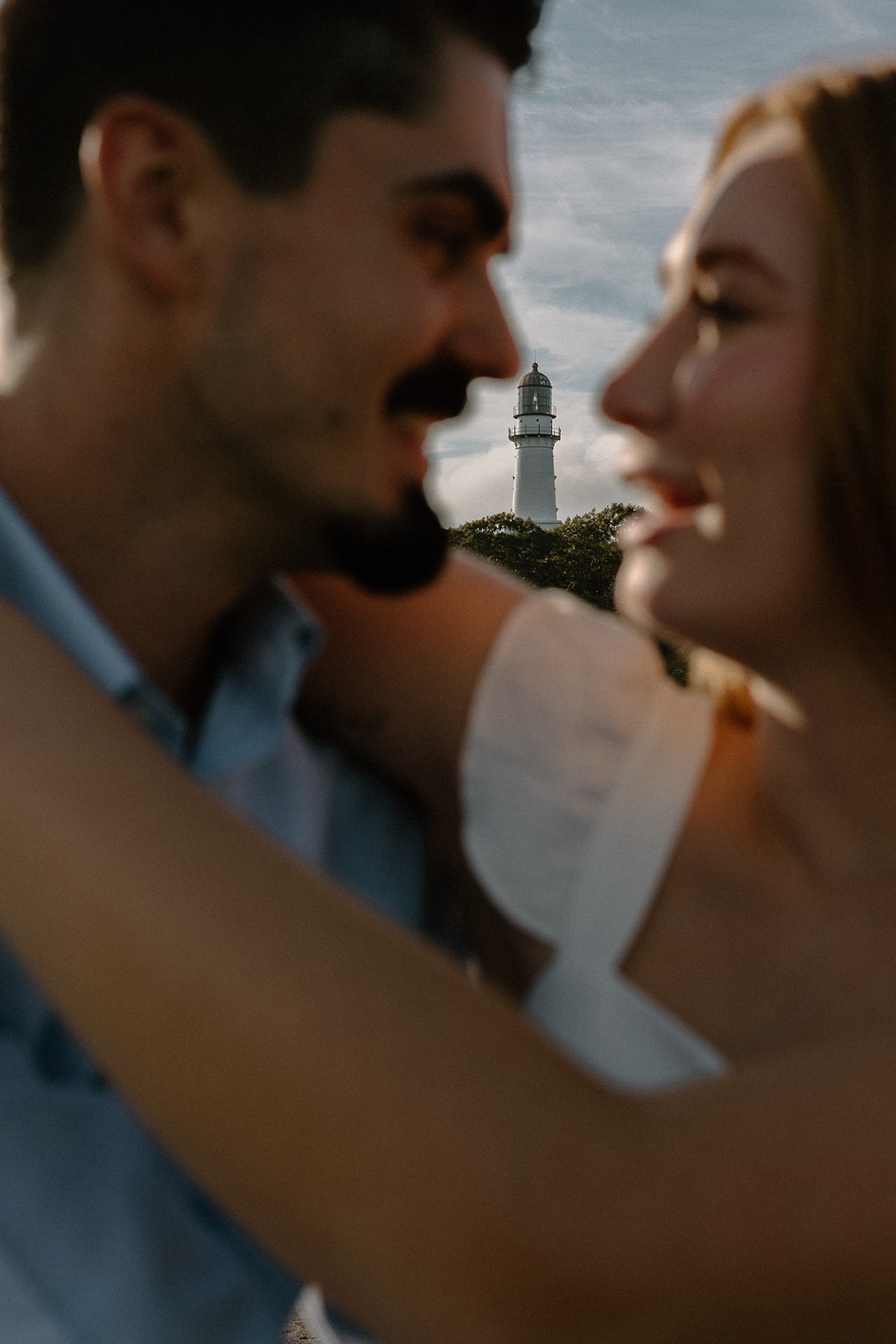 Romantic blurred portrait of a couple embracing with a lighthouse in the distance, showcasing the charm of coastal Engagement Photo Locations.