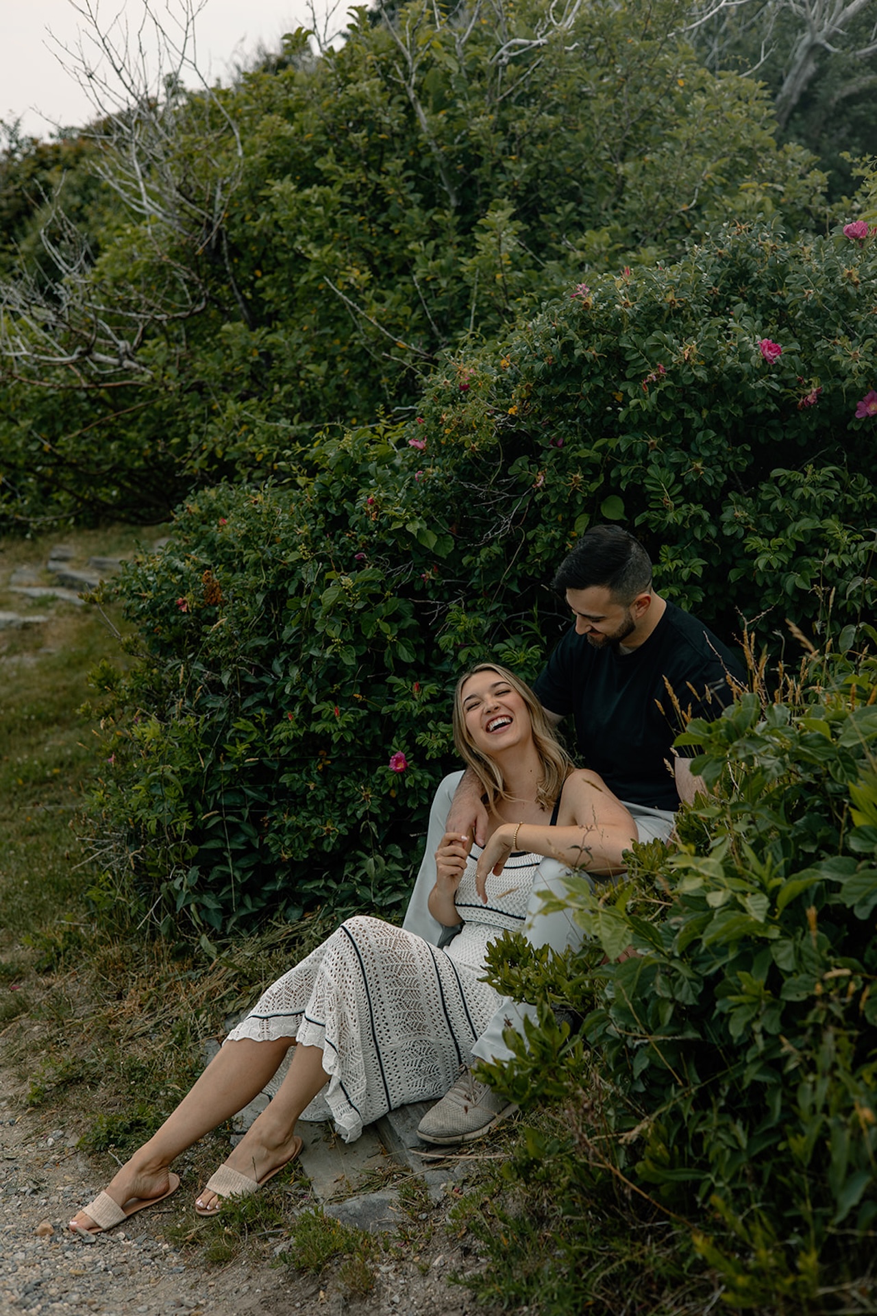 Couple sitting among lush green bushes, laughing together while surrounded by summer blooms.