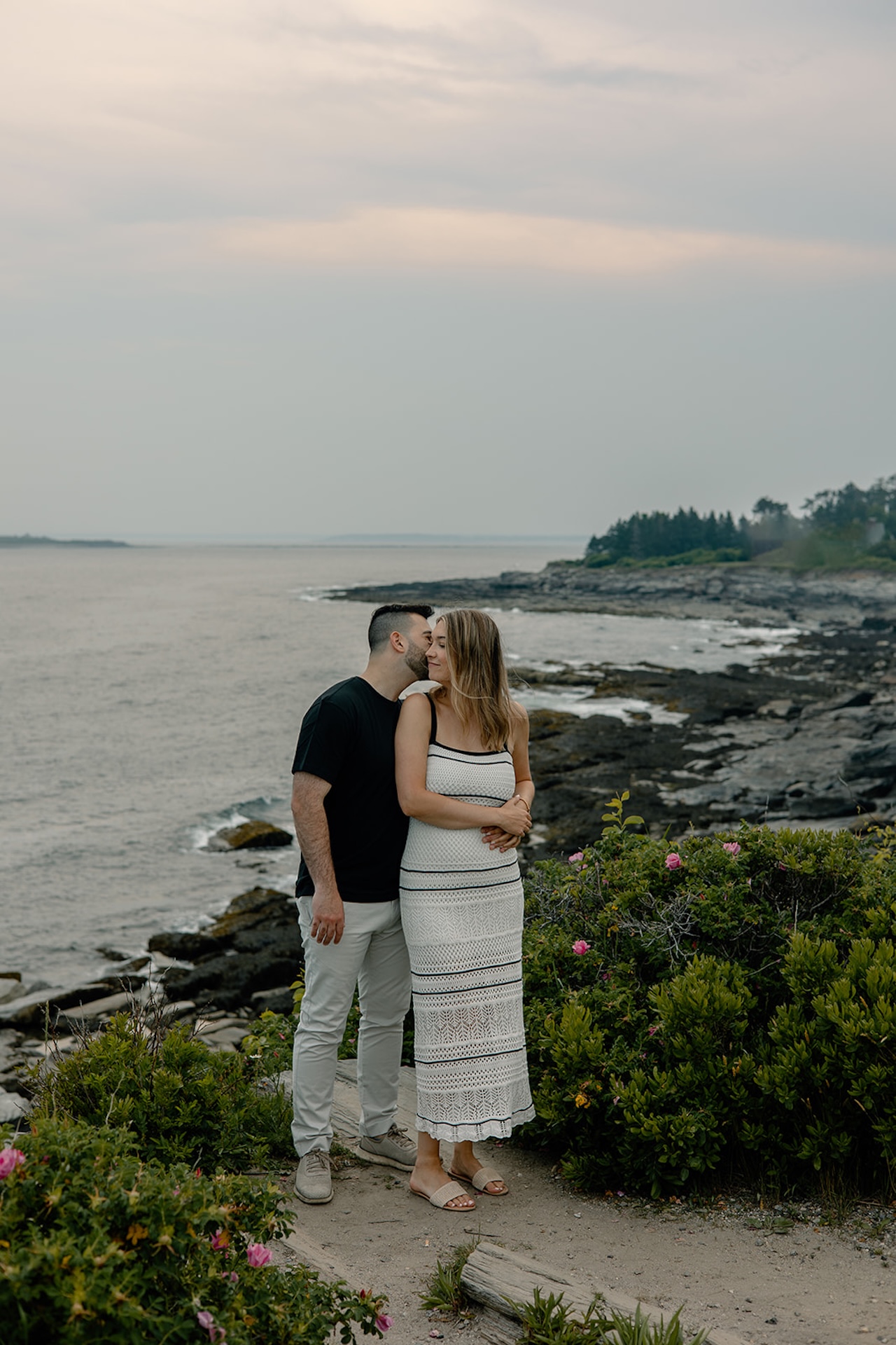 Couple standing on rocky coastline, holding each other with waves behind them at a classic Portland, Maine Engagement Photo Locations setting.