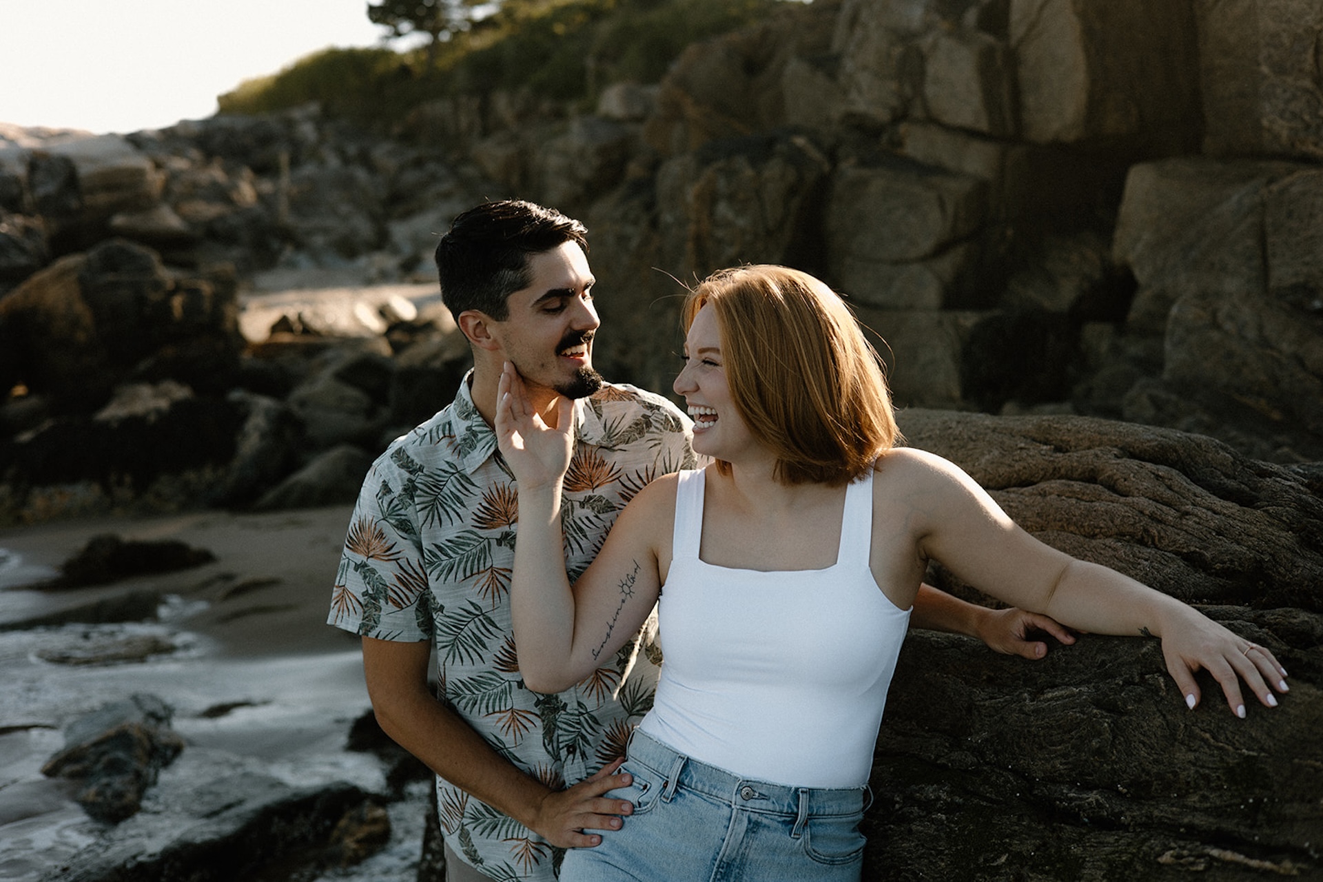 Couple laughing together near seaside rocks, playful energy at one of the prettiest coastal Engagement Photo Locations in Maine.