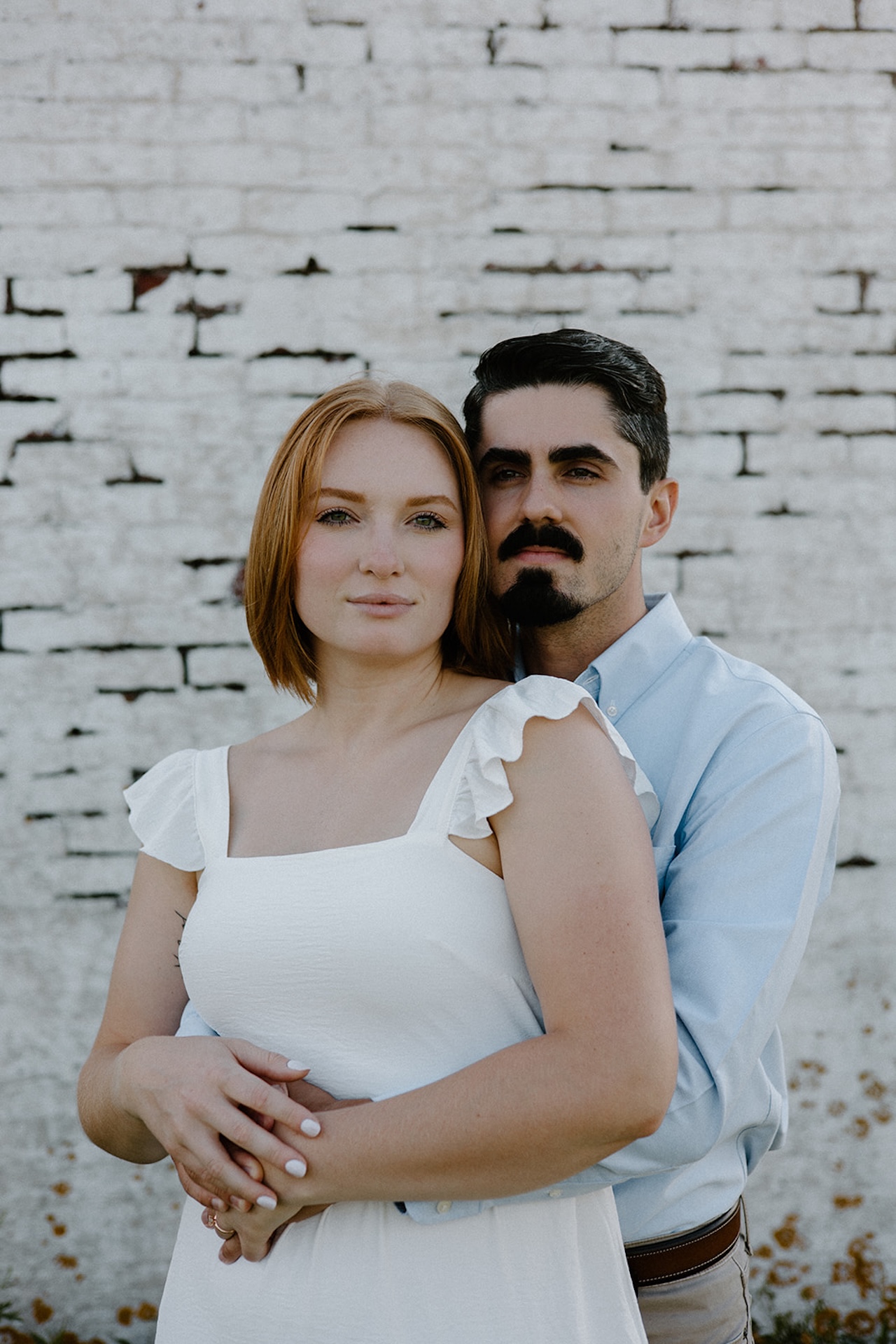 Couple standing in front of a white textured wall, holding each other and looking toward the camera.
