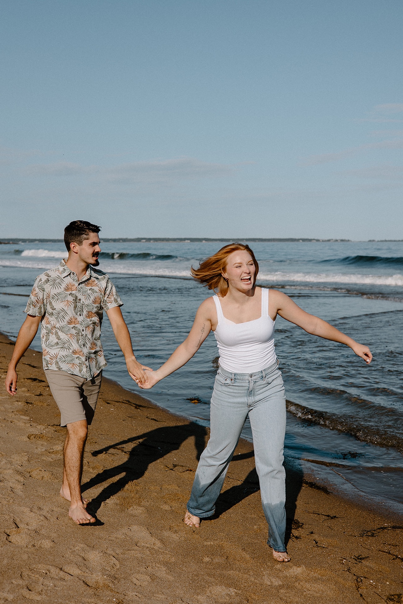 Couple holding hands and laughing while walking barefoot along the beach, capturing carefree love and natural connection.