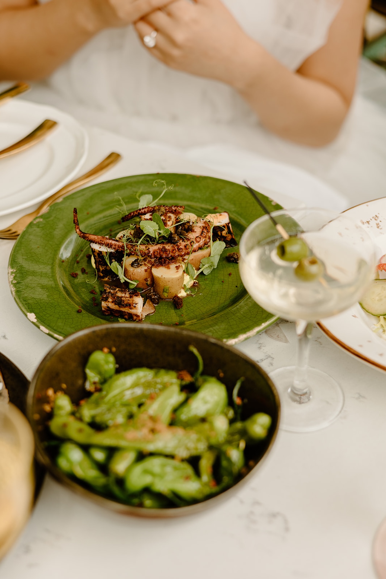 Elegant flat lay of a meal with grilled octopus, shishito peppers, and a martini on the table.