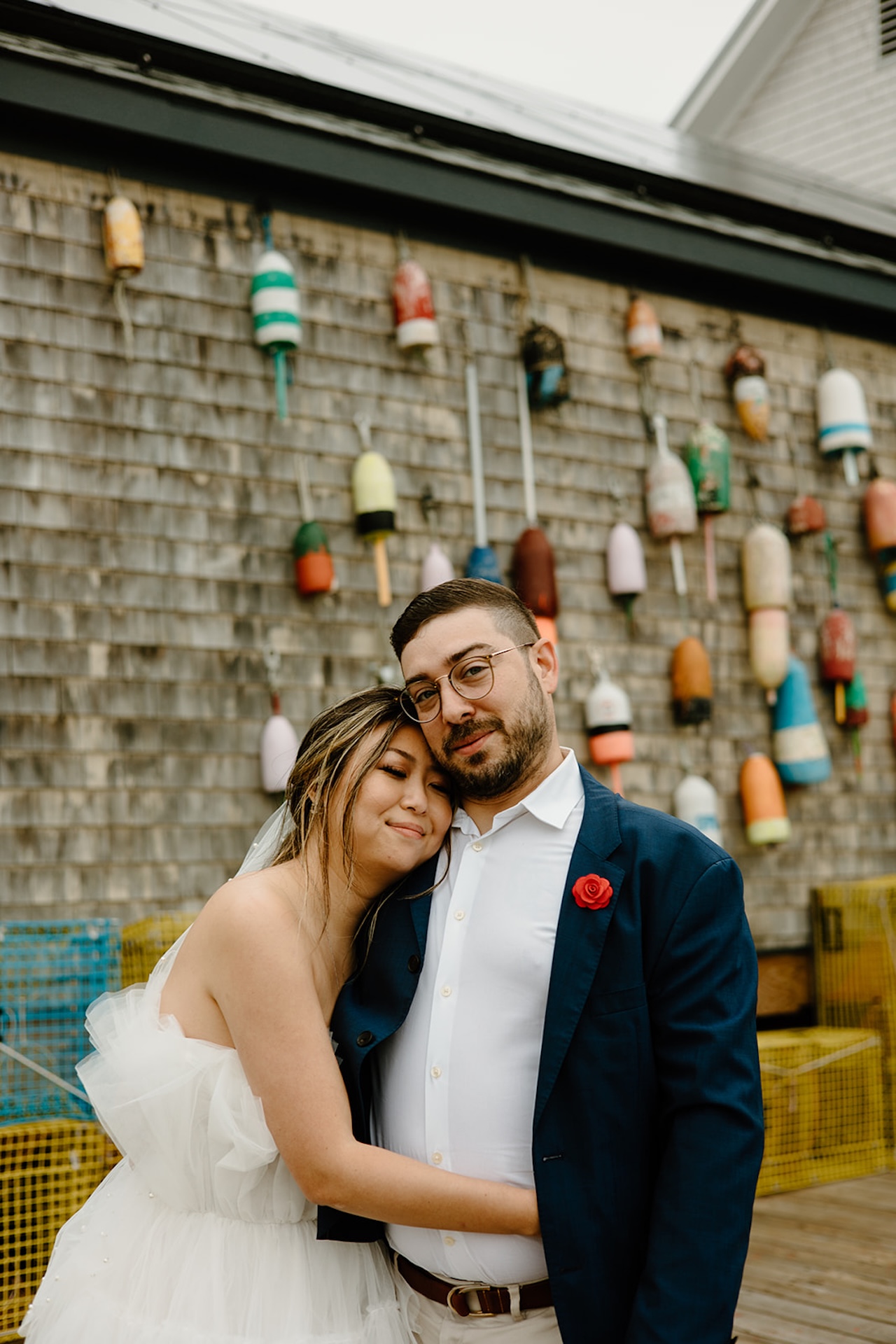 Bride leaning on groom’s shoulder in front of a wall covered with colorful buoys at one of Maine’s seaside Engagement Photo Locations.
