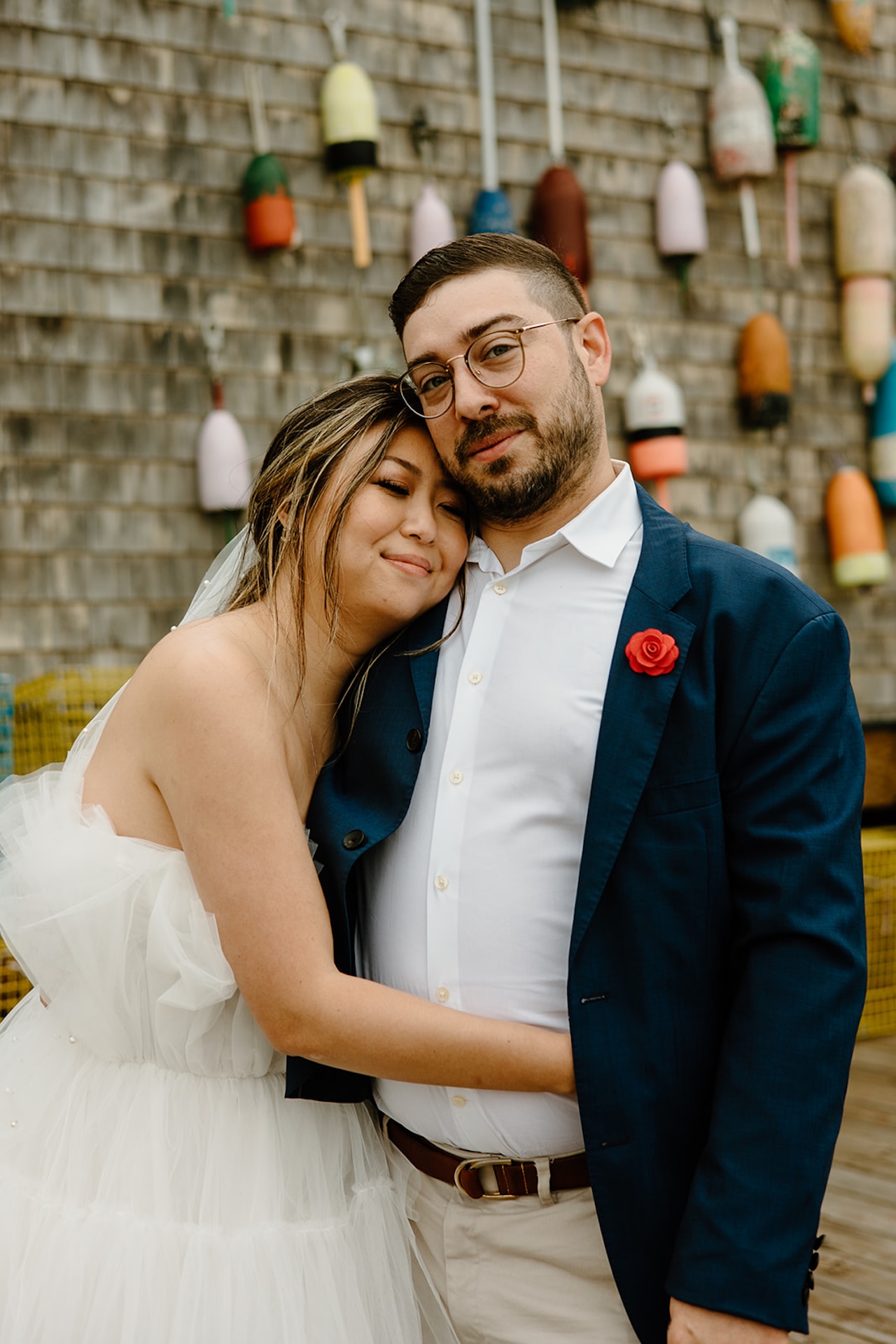 Bride and groom embracing with soft smiles against a wall of colorful buoys, capturing a cozy, coastal Maine wedding moment for spring engagement photo locations near Portland Maine.