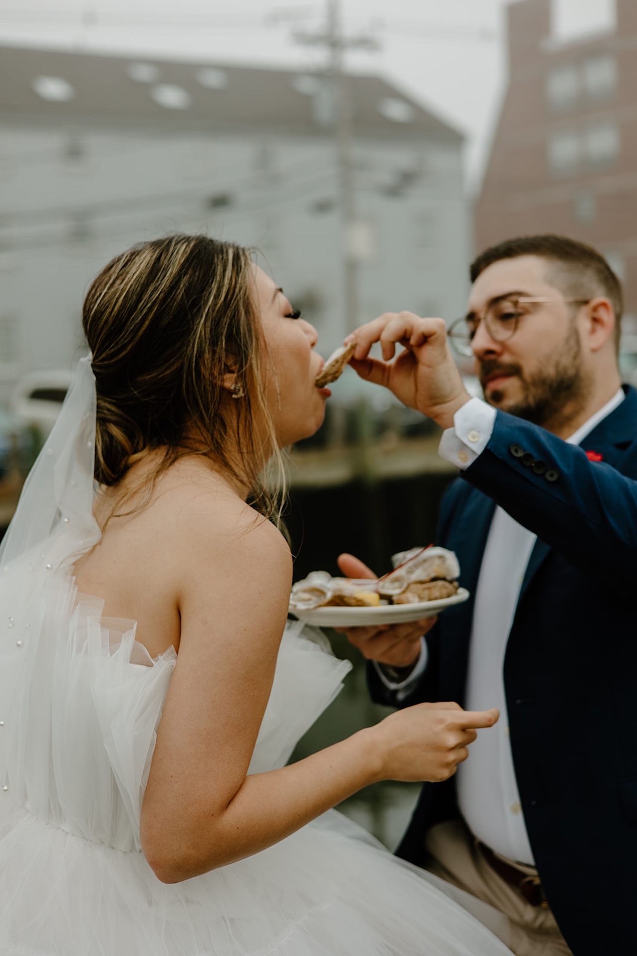 Groom feeding his bride oysters by the harbor, capturing a playful and candid wedding moment at the waterfront.
