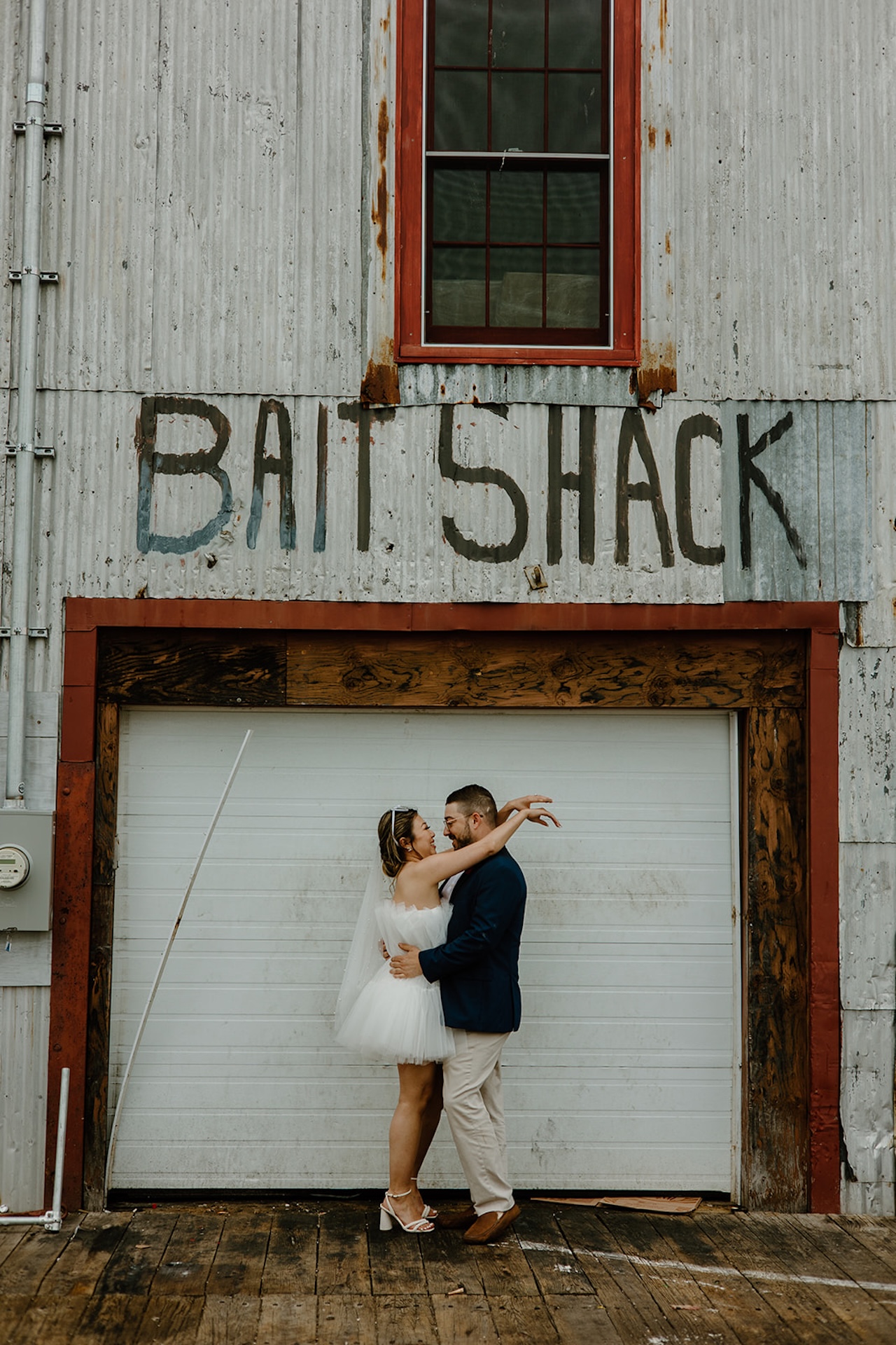 Bride and groom embracing in front of a rustic building that reads “Bait Shack,” blending modern style with coastal charm.
