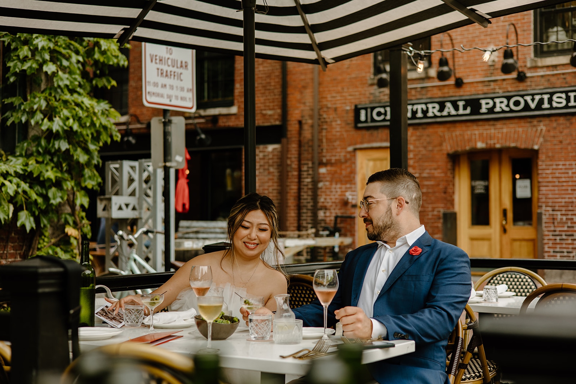 Bride and groom enjoying drinks together under a striped umbrella outside a restaurant downtown.