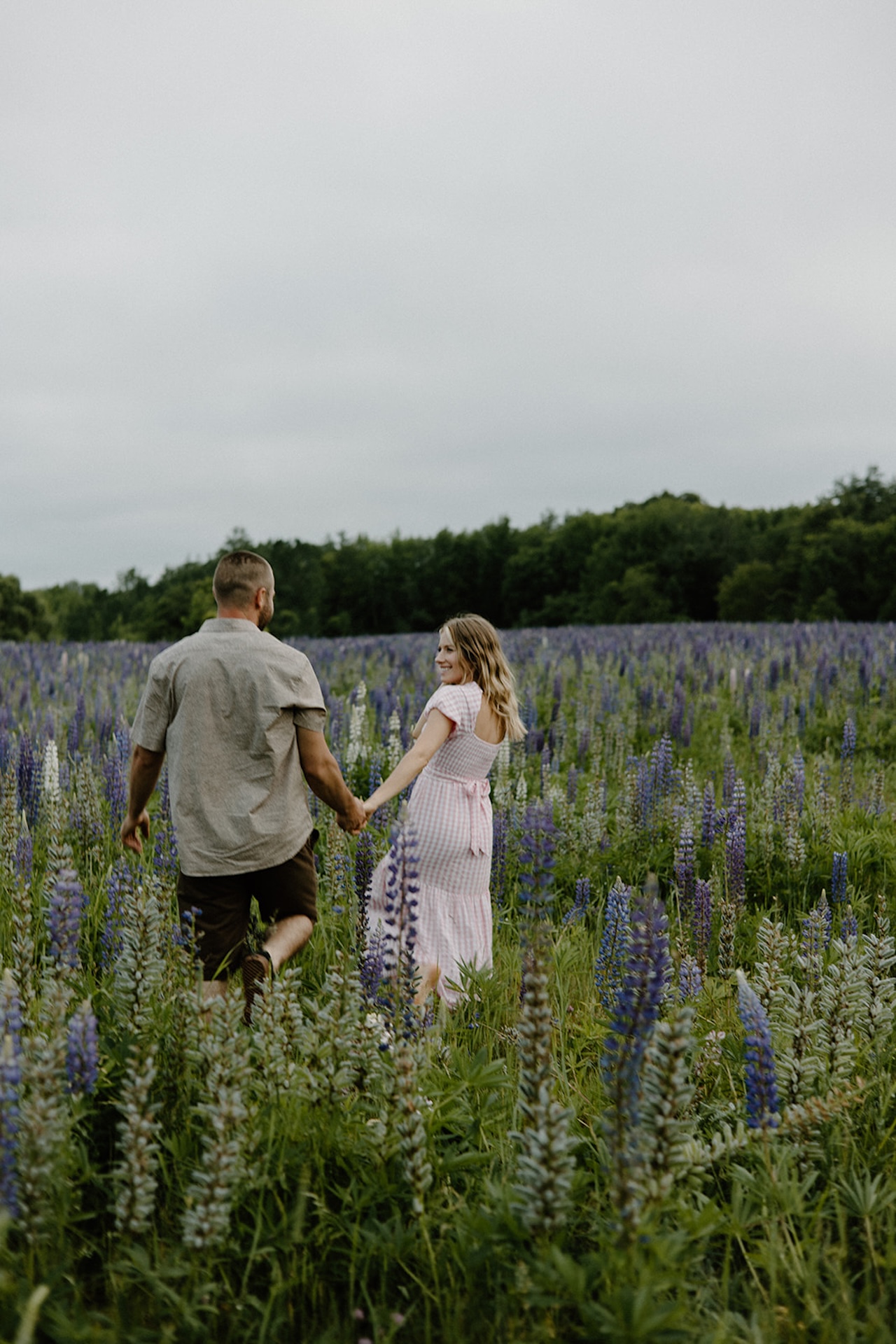 Couple walking hand in hand through a wildflower field, smiling as they look back toward the camera.