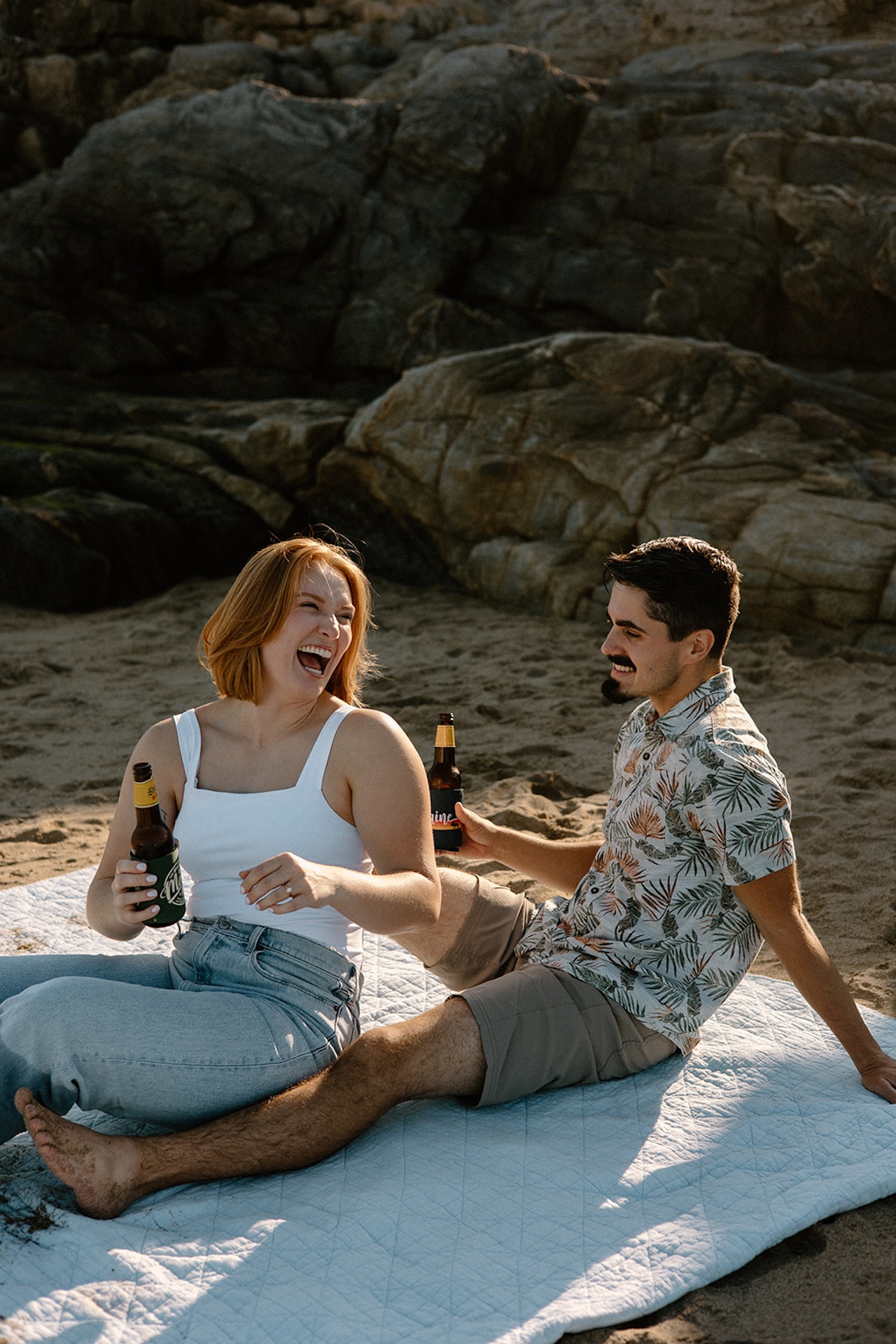 Couple laughing together on the sand, holding drinks and soaking up the sun at coastal Engagement Photo Locations.
