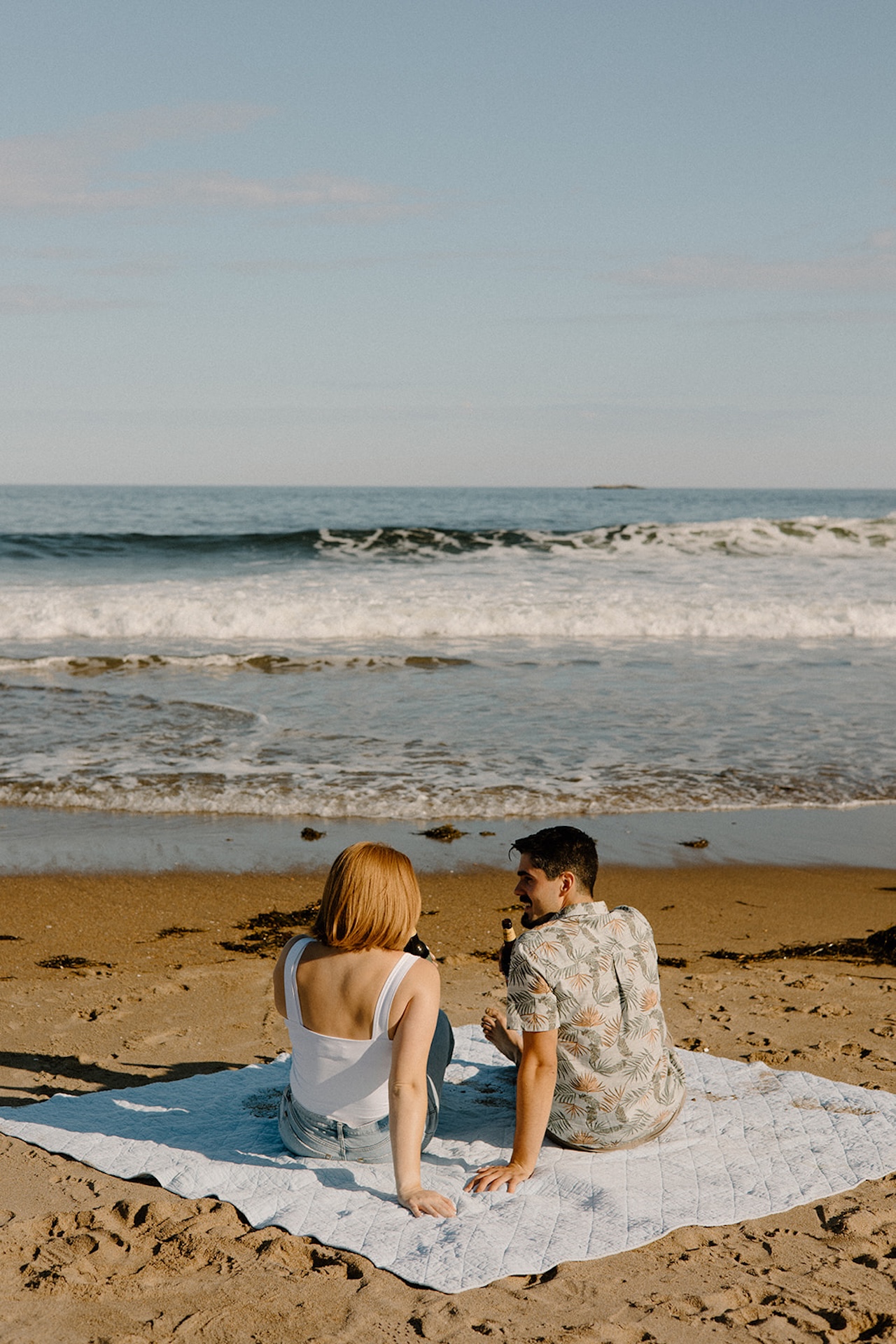 Couple sitting on a blanket at the beach, looking toward the ocean waves under a clear sky.
