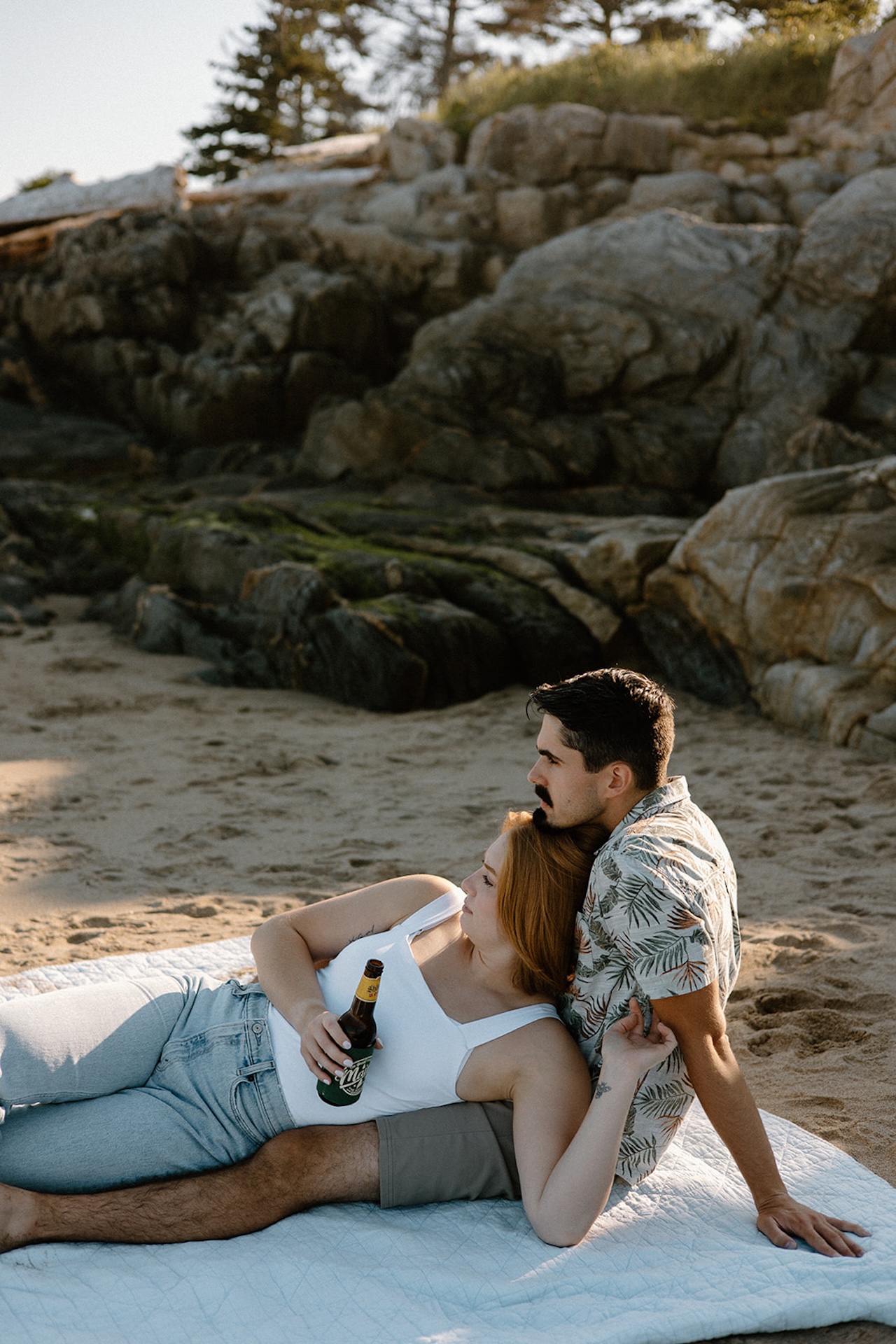 Couple sitting on a blanket at the beach, sharing drinks and leaning against each other as the sun sets.