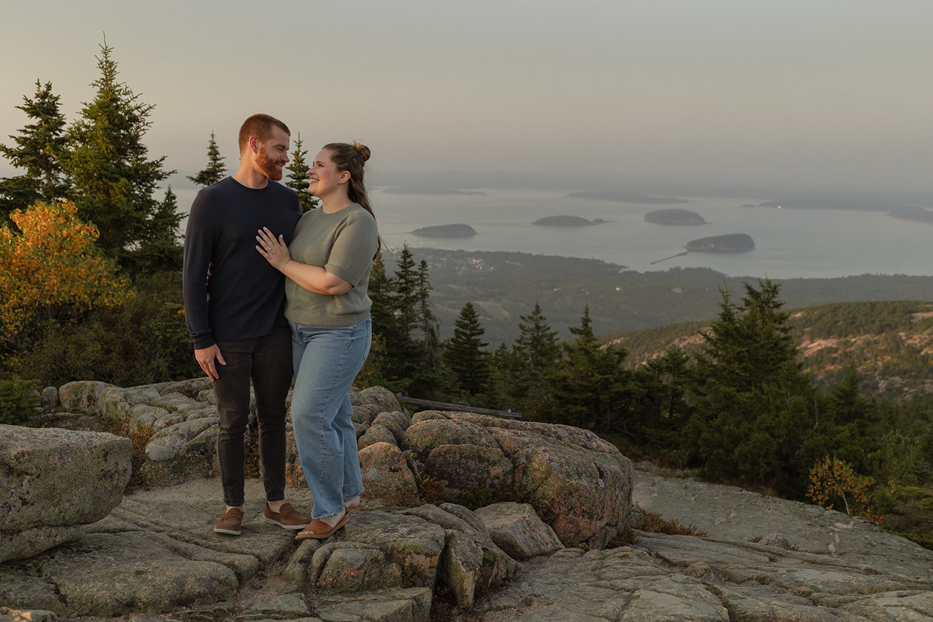 A couple smiling at each other on top of a mountain at golden hour, with the sweeping coastal landscape behind them, captured as Acadia engagement photos.