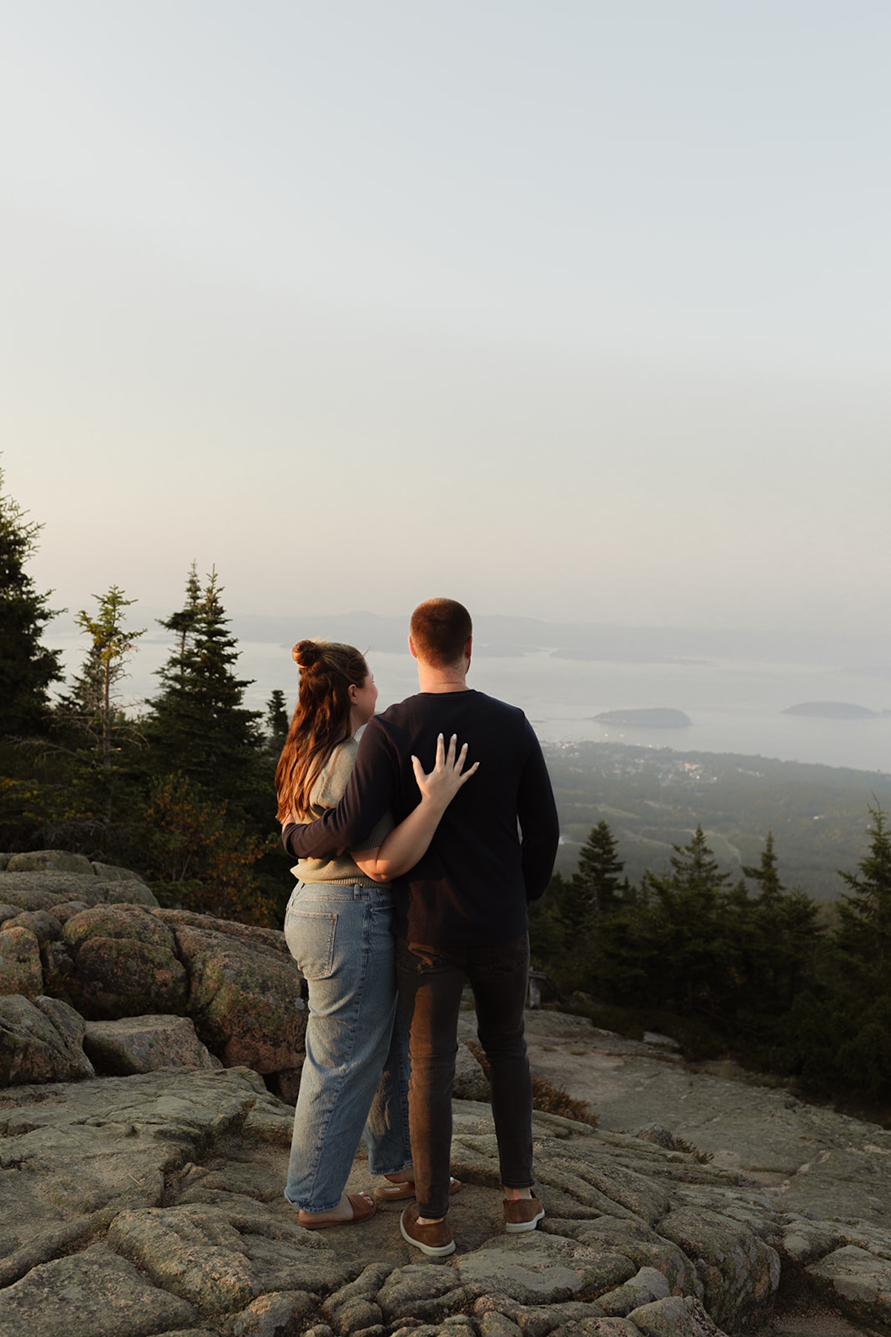 A couple standing on a rocky overlook, arms wrapped around each other while gazing out toward the distant islands and hazy coastline.