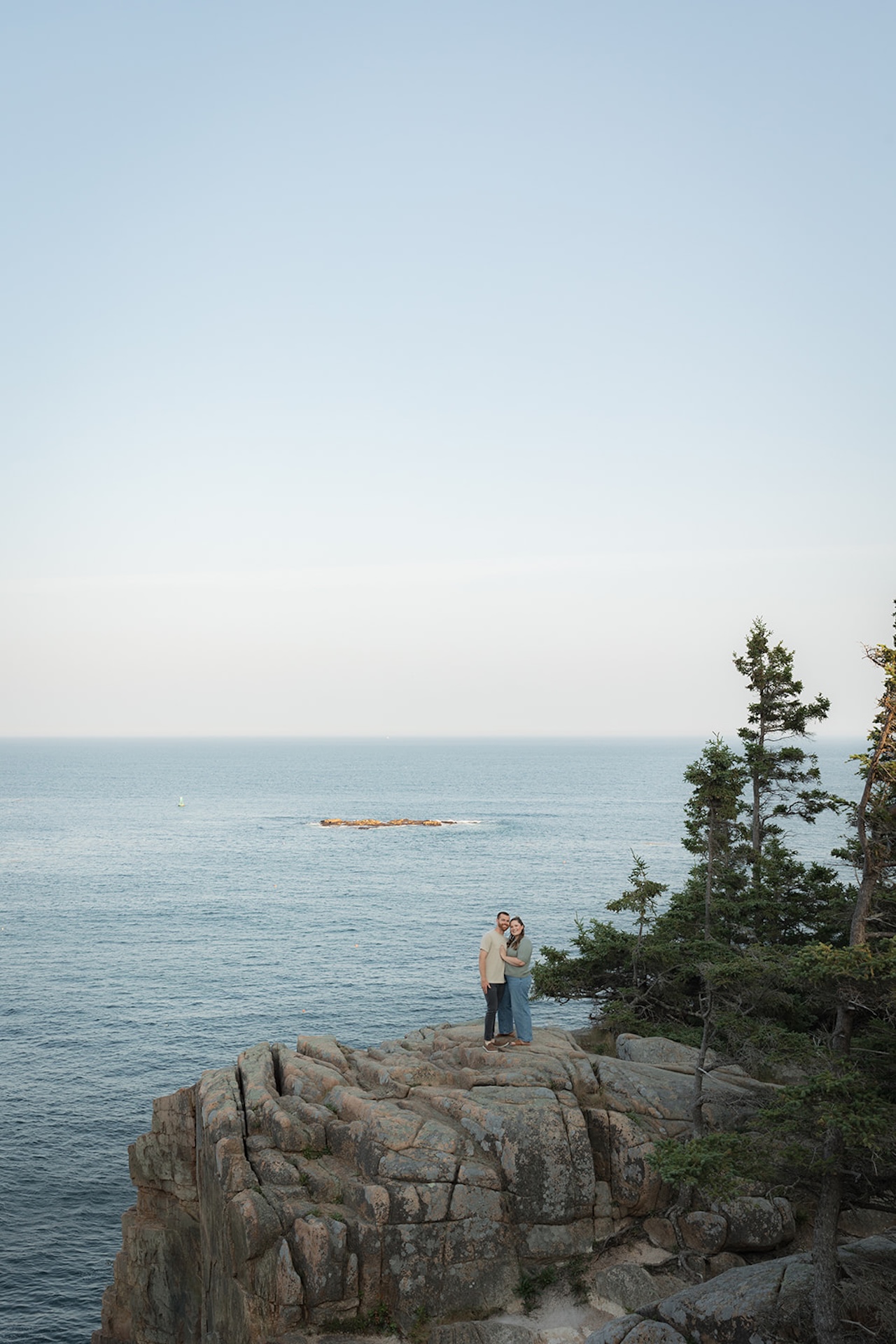 The couple standing on a rocky cliff at Acadia National Park with the water in the back ground.