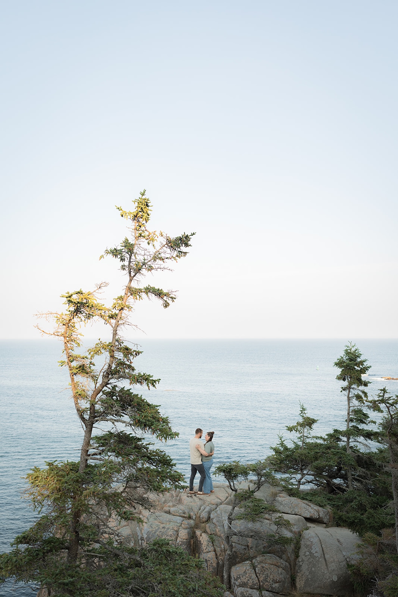 A couple hugging on a rocky cliff edge with the blue ocean stretching out behind them, framed softly by nearby branches.