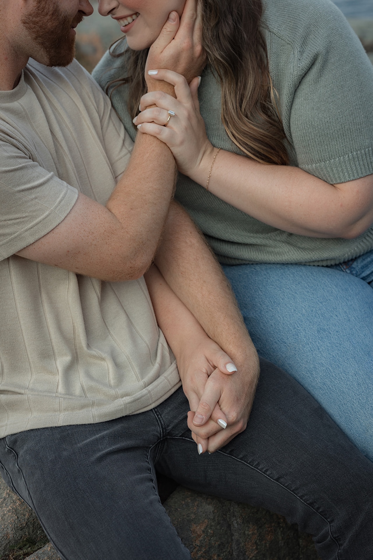 A close, intimate shot of a couple holding hands and leaning in, showing their rings and soft smiles as they sit together on the rocks.