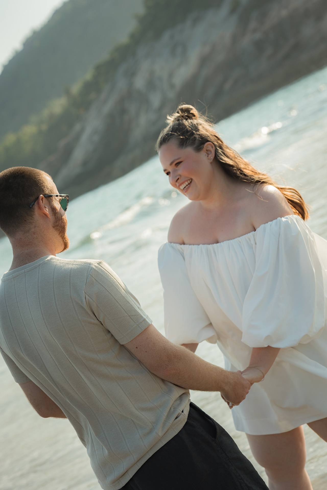 A candid shot of a couple laughing while holding hands at the shoreline, waves and cliffs behind them as the sun hits the water.