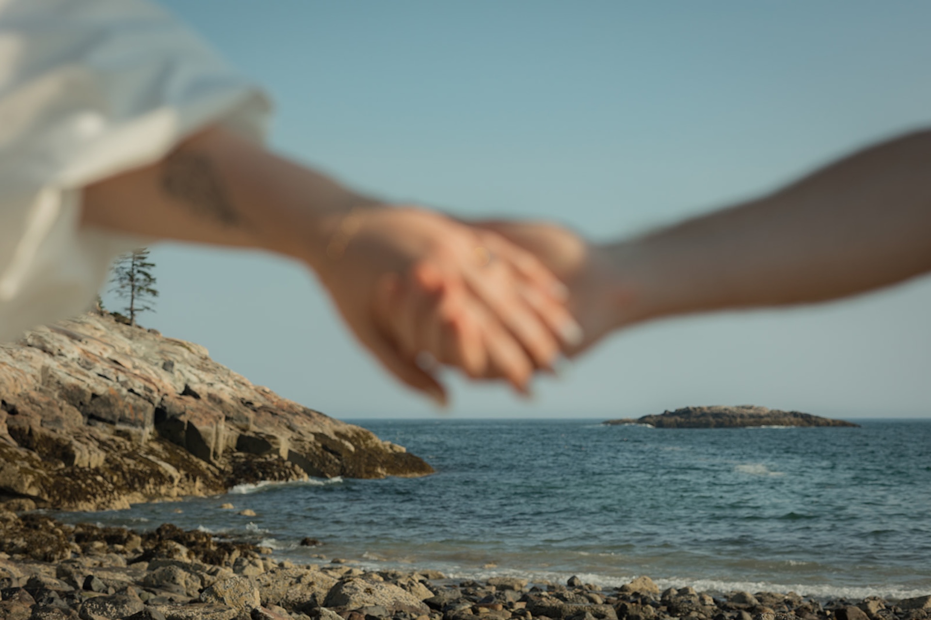 The couples hands in the frame out of focus with the beach at Acadia National Park in focus behind them.