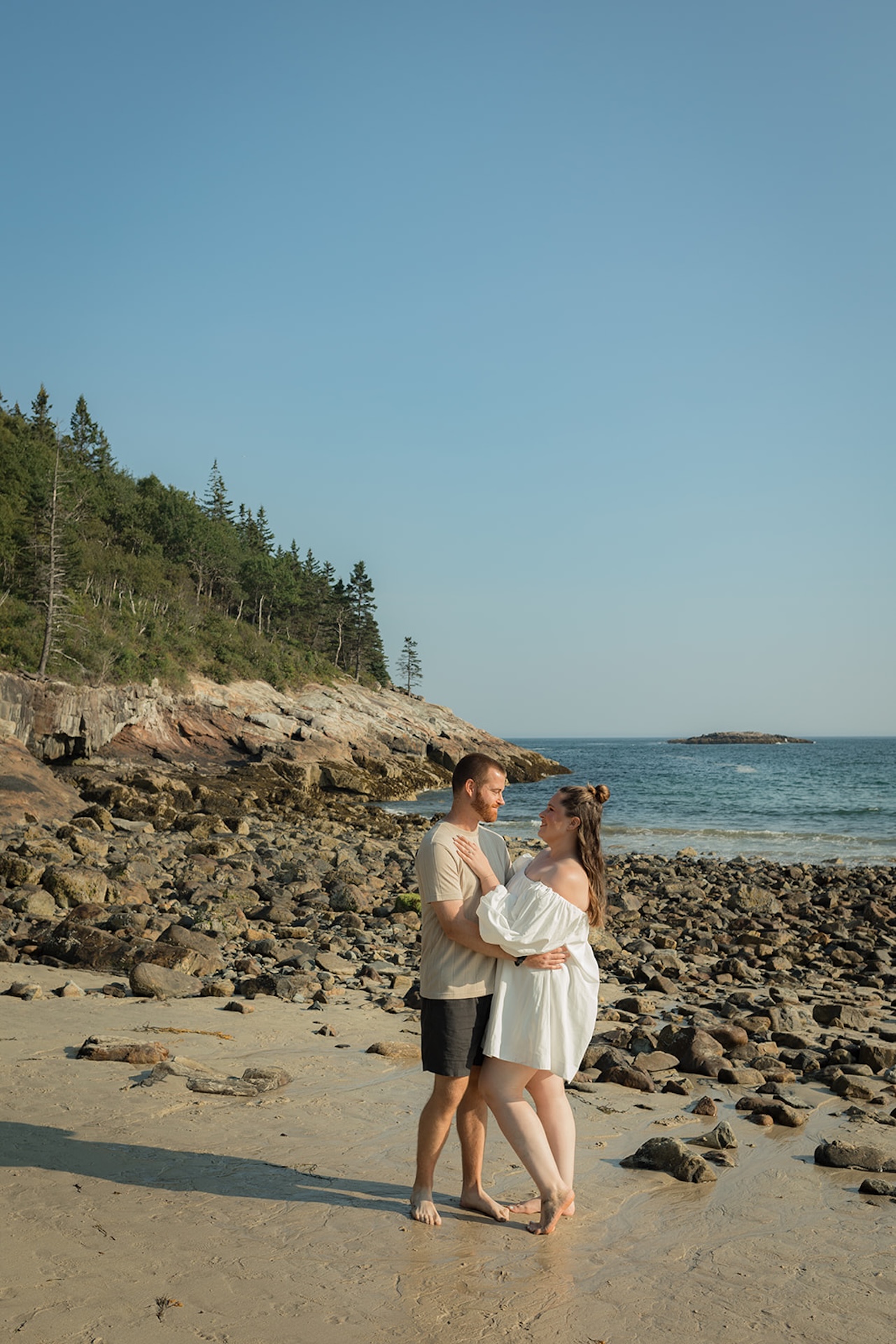 A couple standing together on a rocky beach with the ocean behind them, smiling at each other as the sunlight hits the shoreline, captured as Acadia engagement photos.
