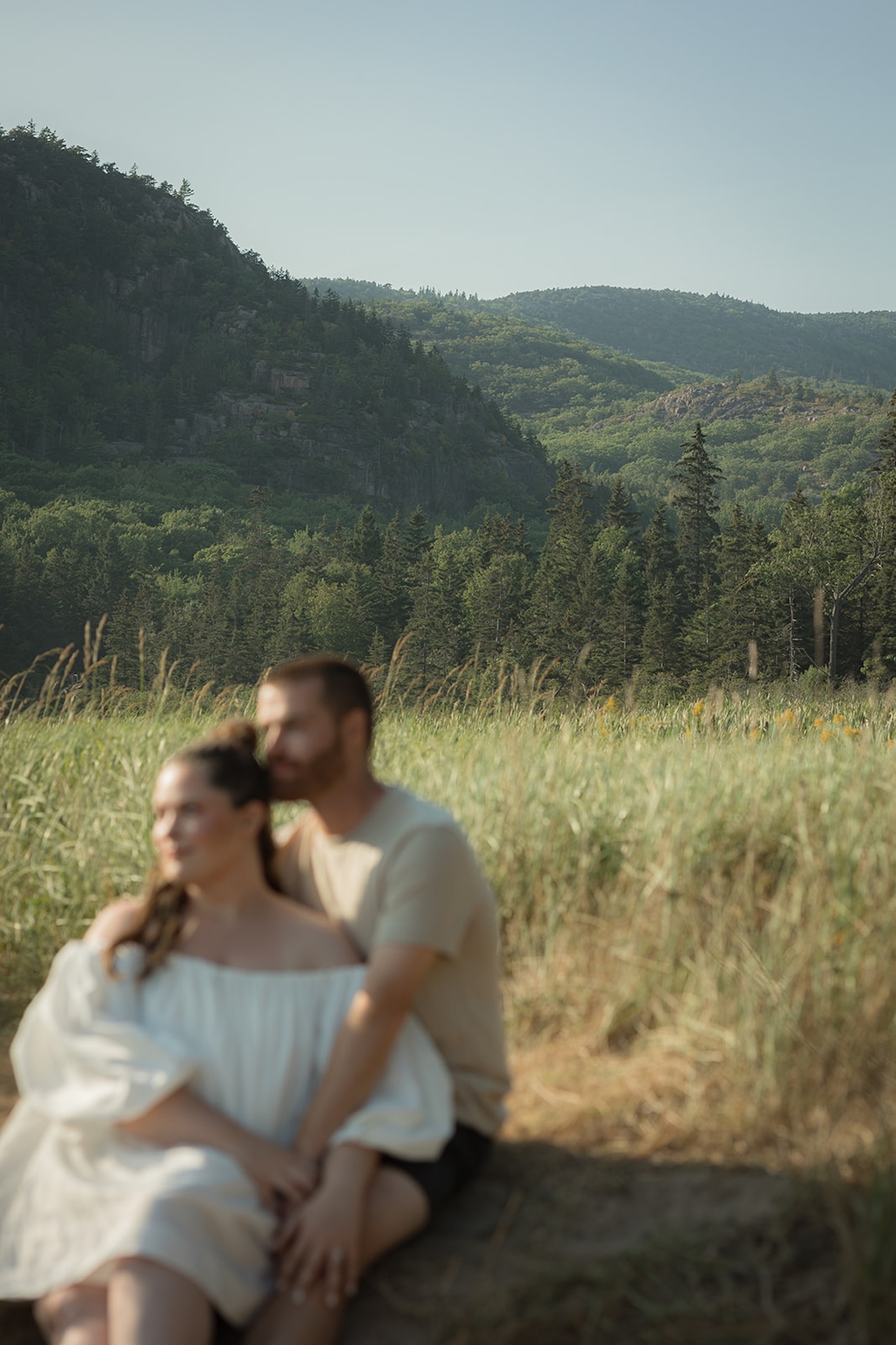 Couple sitting in a grassy field with views of Acadia mountains in the background during their Acadia national park engagement photos
