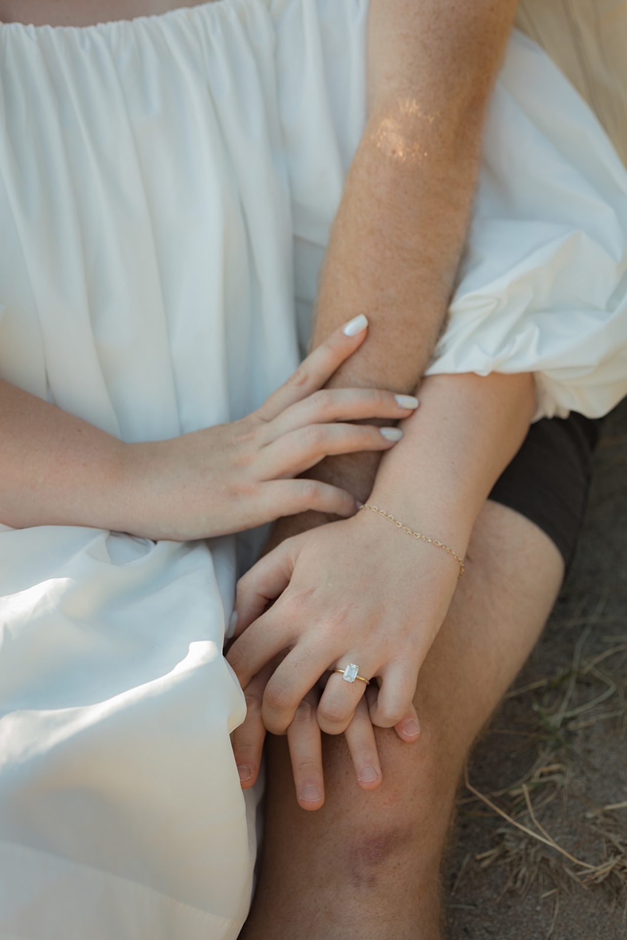 A close-up of intertwined hands resting gently on a partner’s knee, highlighting the engagement ring and tender touch as they sit together outdoors.