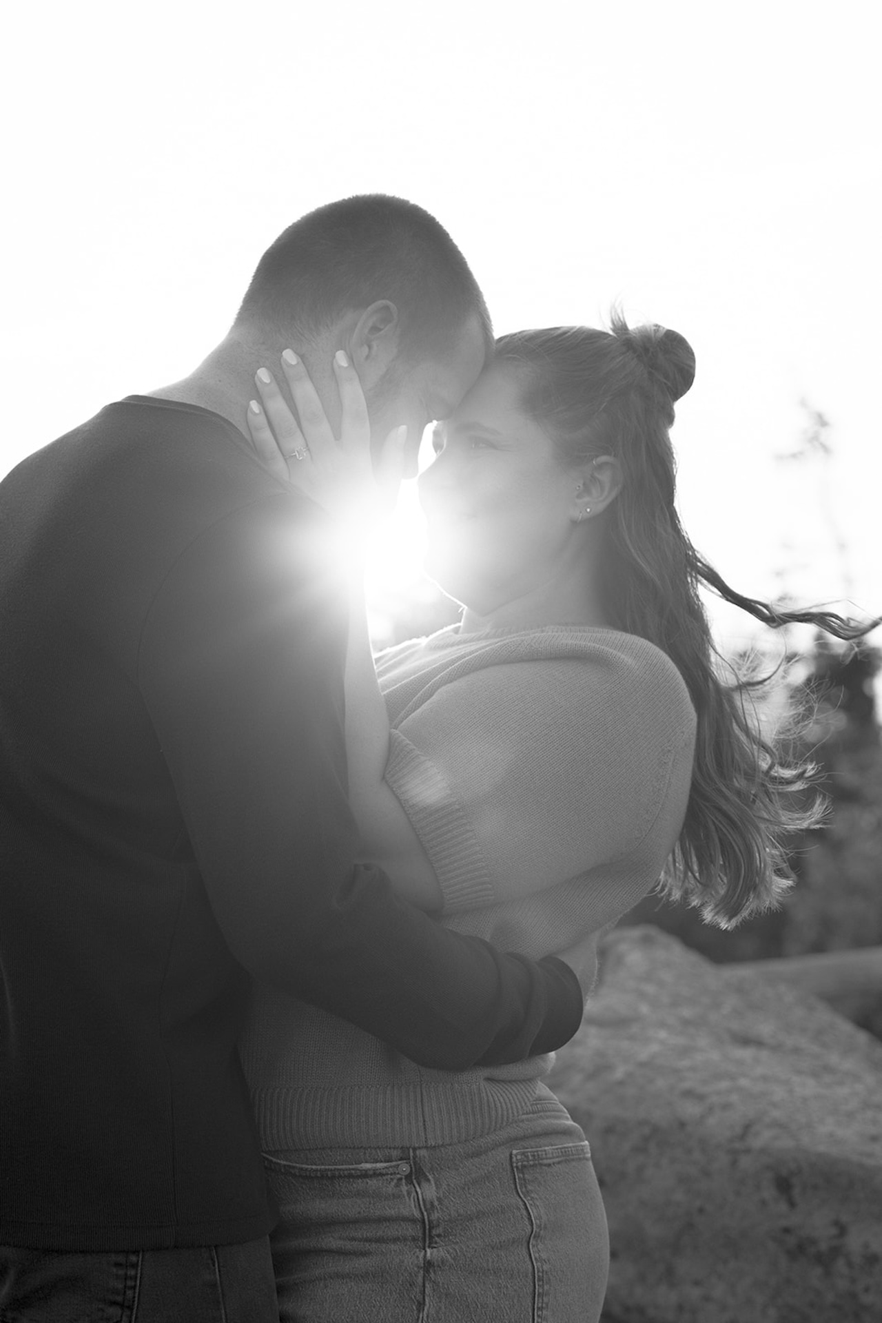 A black-and-white photo of a couple holding each other with sunlight flaring between them, captured during their Acadia engagement photos.