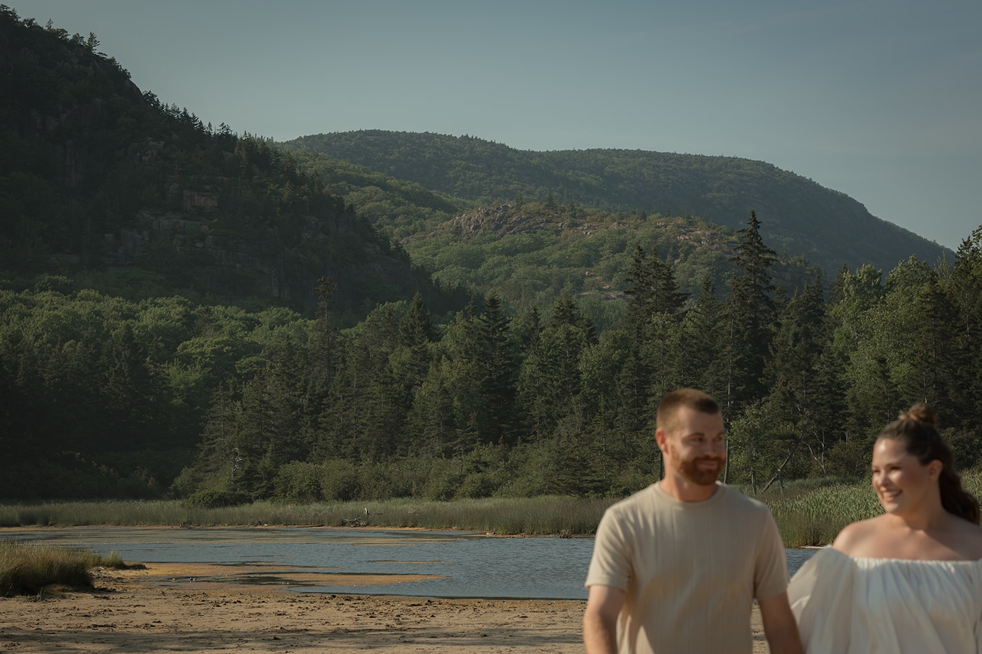 A blurred, candid moment of a couple walking along a sandy shoreline with forested mountains rising in the background.