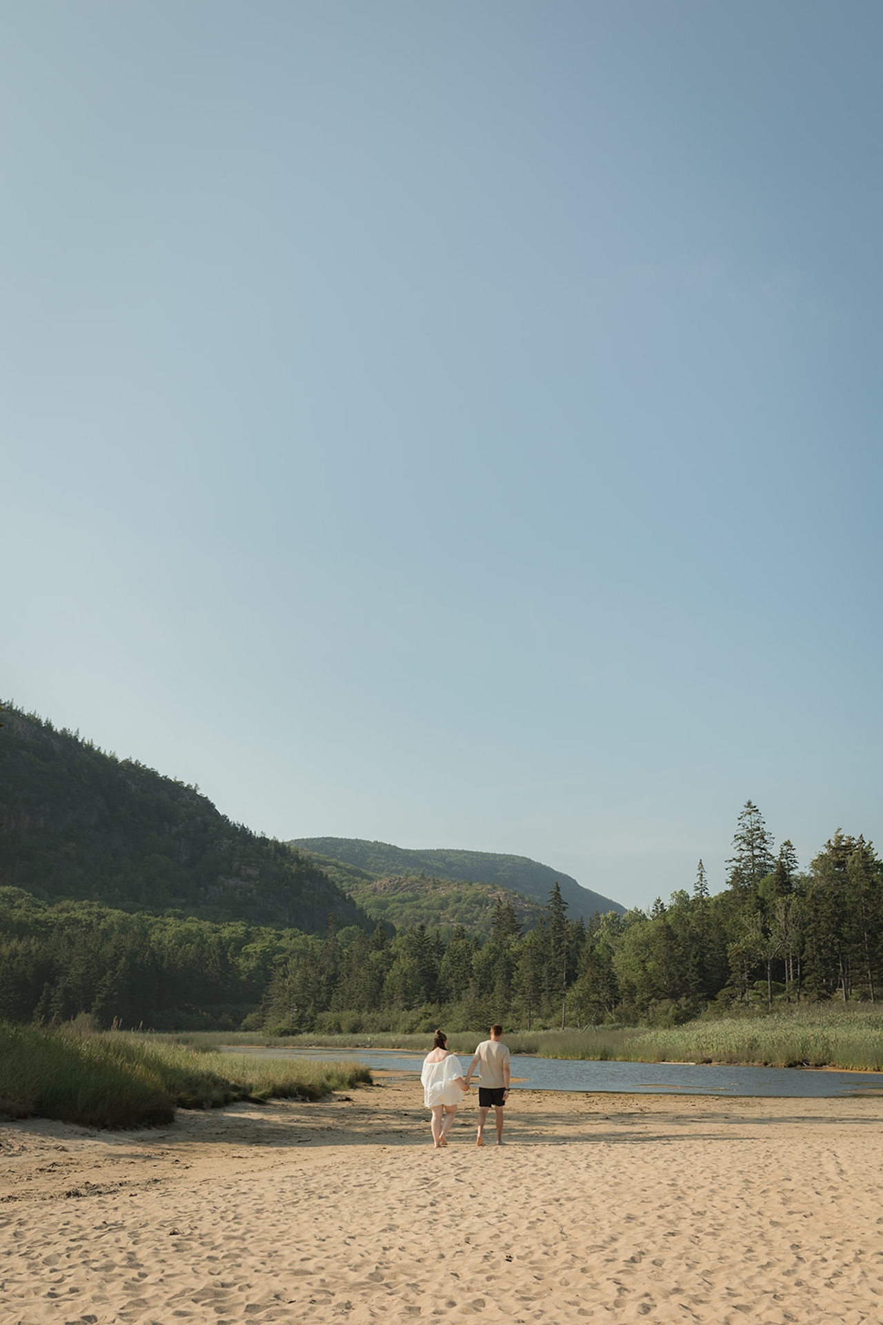 A wide landscape image of a couple walking hand-in-hand across sandy terrain toward a quiet river, surrounded by mountains and evergreens, photographed as Acadia engagement photos.