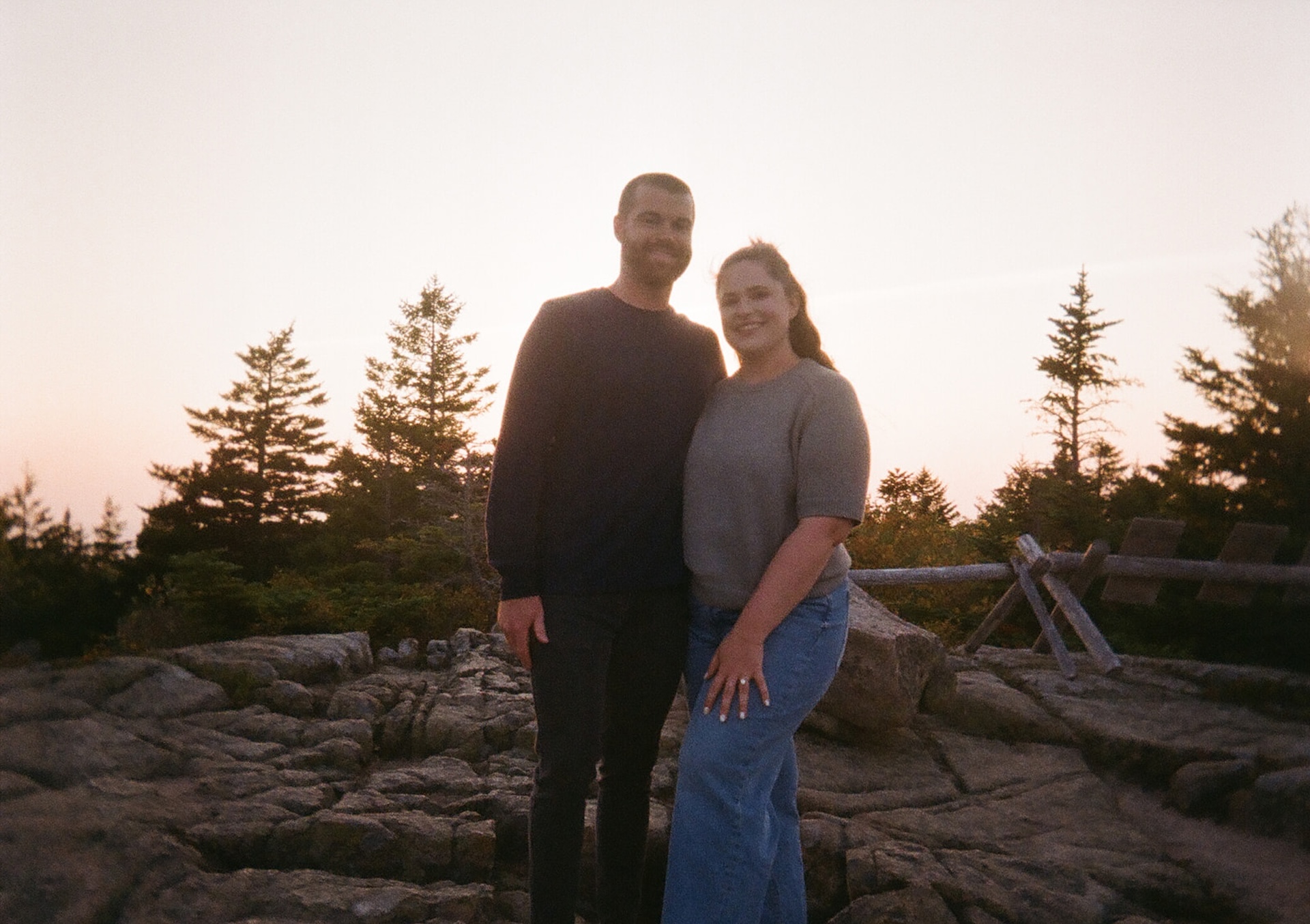 A film-style image of a couple standing together on a rocky overlook at sunset, smiling with the sweeping coastal view behind them as part of their Acadia engagement photos.