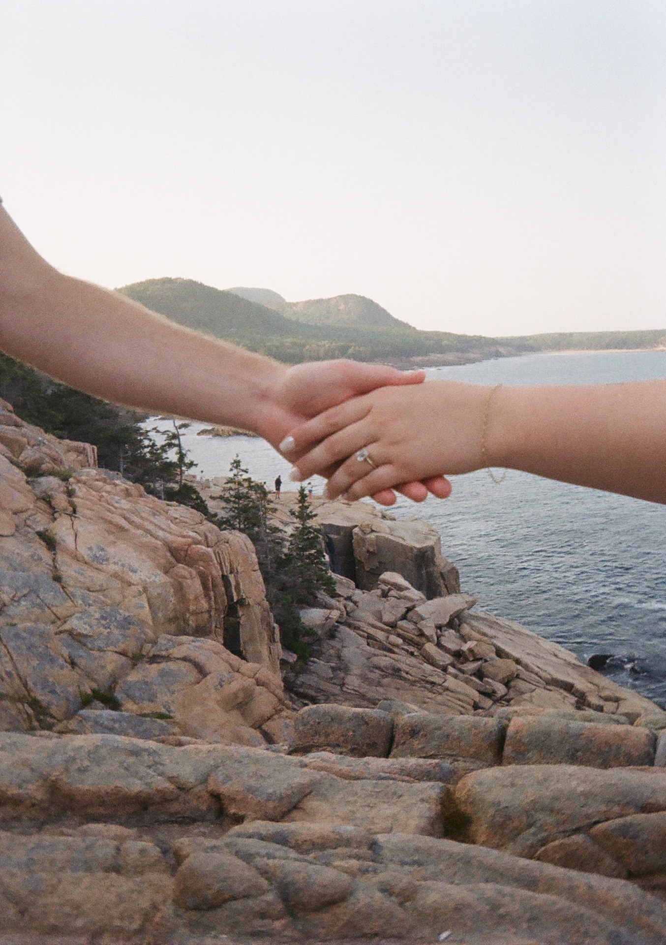 A detail shot of a couple holding hands with the rocky cliffs of Acadia National Park in the background