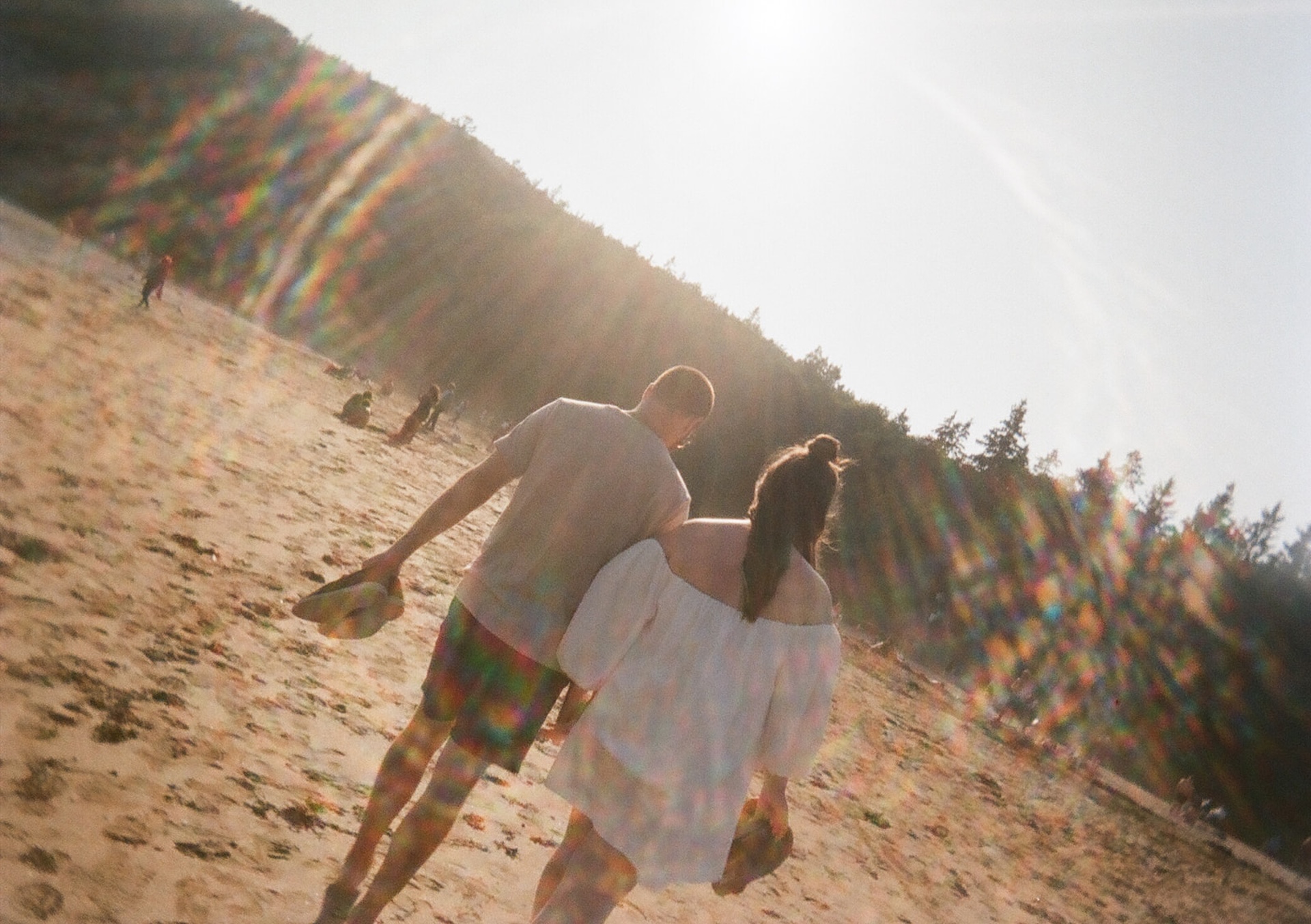 A warm, sun-soaked film photograph of a couple walking across a beach, rainbow flares streaking across the frame in the afternoon light.