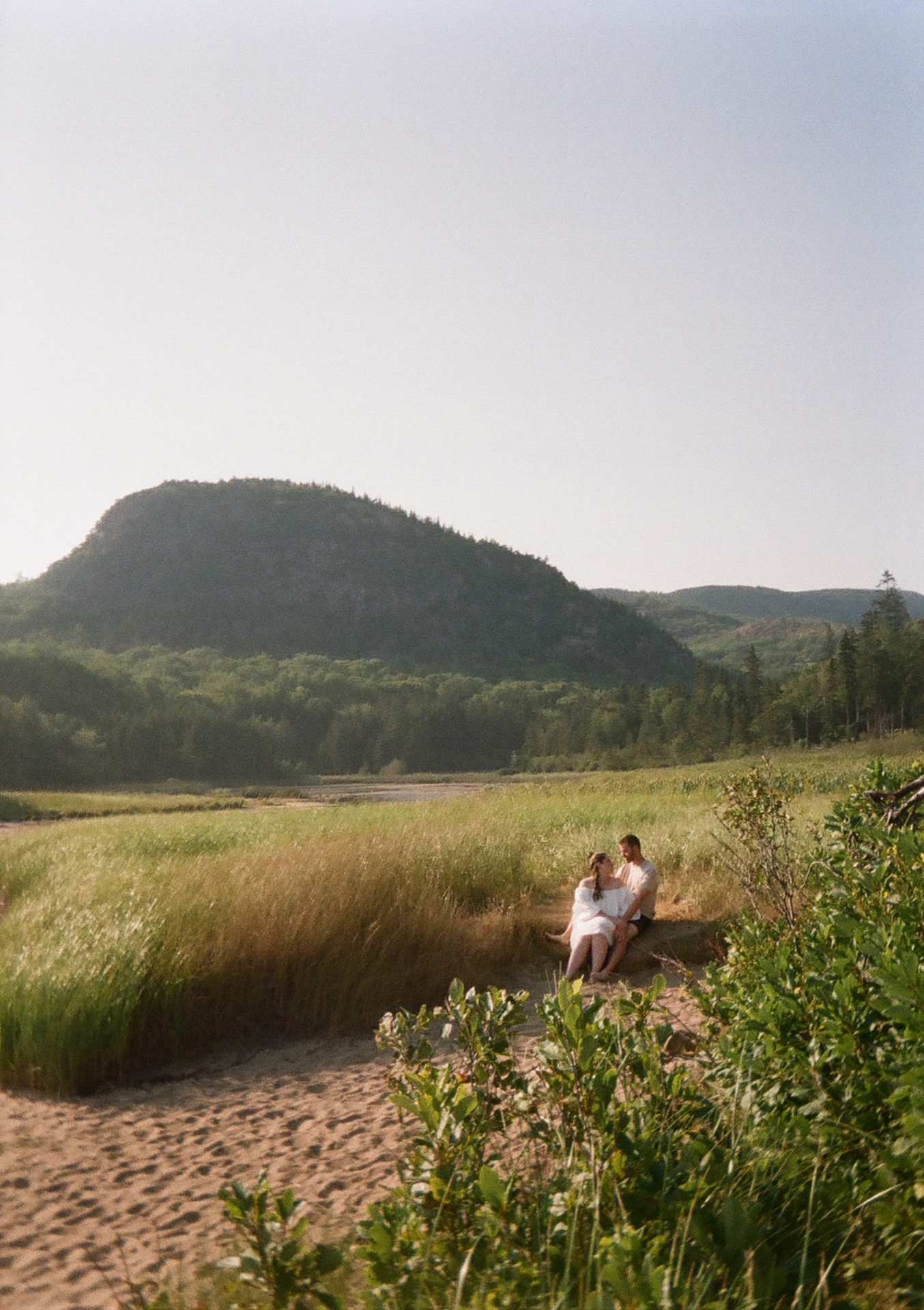 Couple sitting in a grassy field with the view of Acadia mountains in the background during their Acadia national park engagement photos