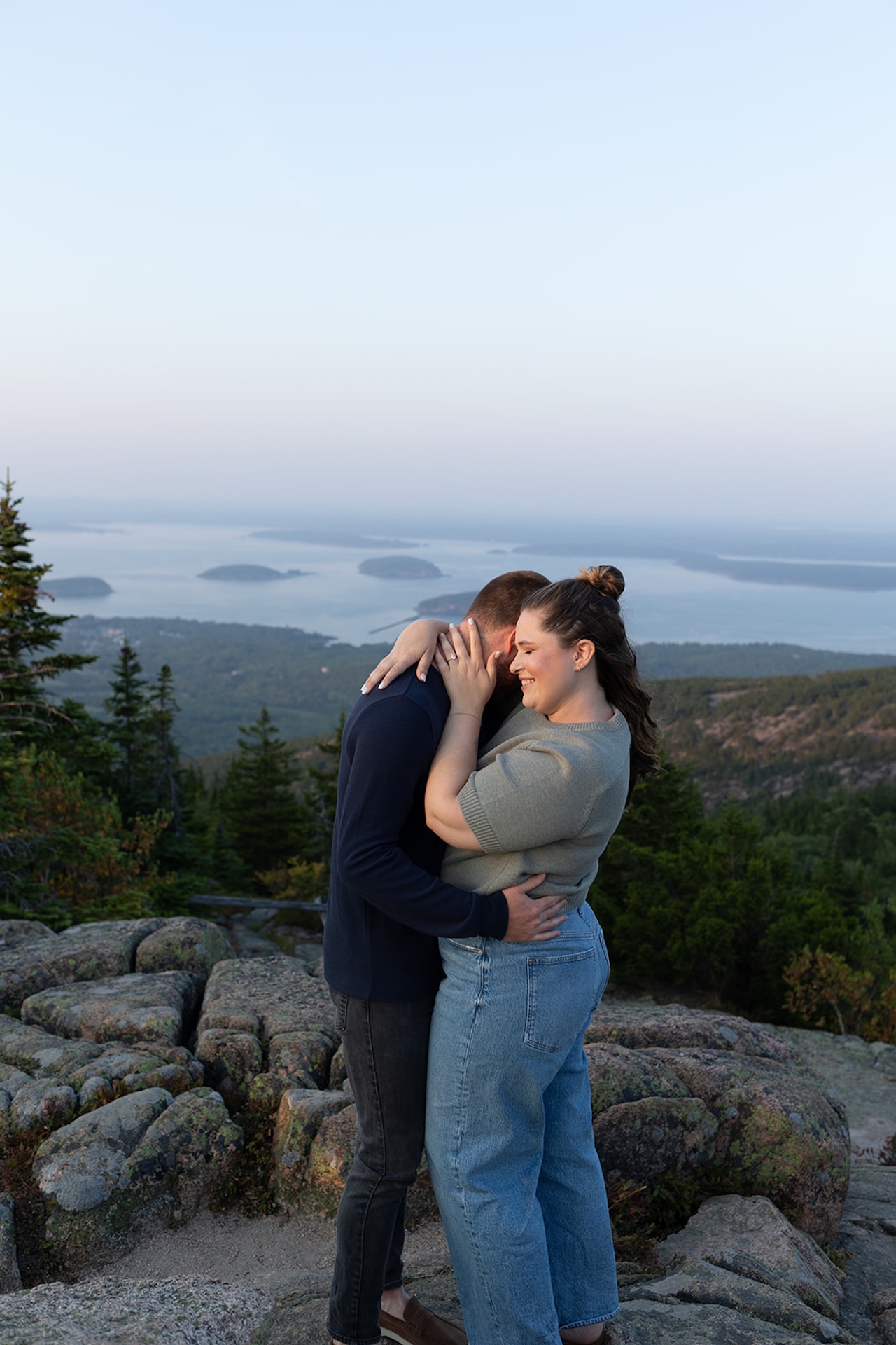 A couple embracing on a rocky mountain overlook at sunset, with distant islands and soft pastel skies behind them, captured as Acadia engagement photos.