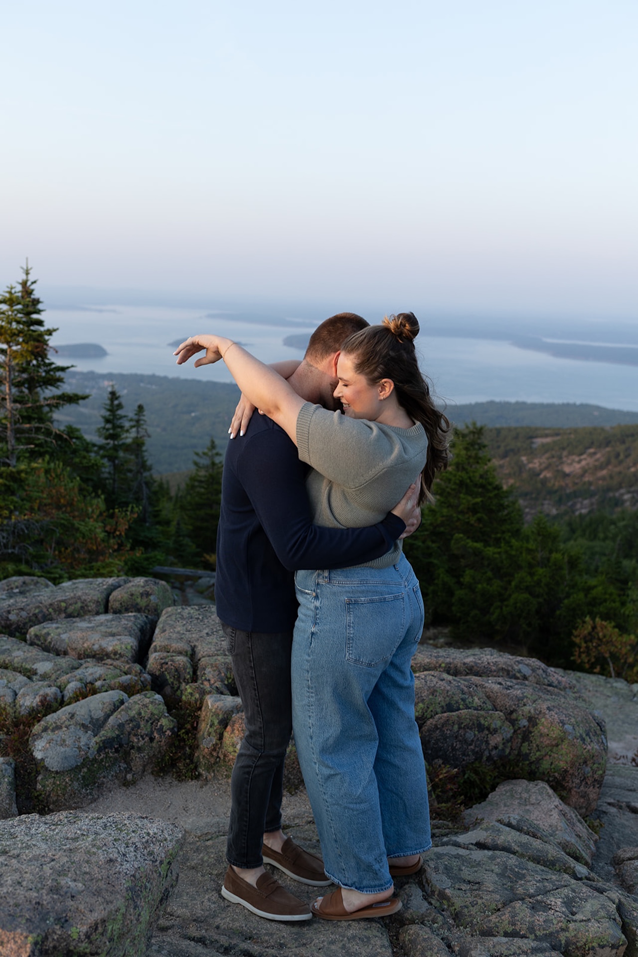 A couple hugging on a rocky mountain overlook at sunset, with distant islands and soft pastel skies behind them, captured as Acadia engagement photos.