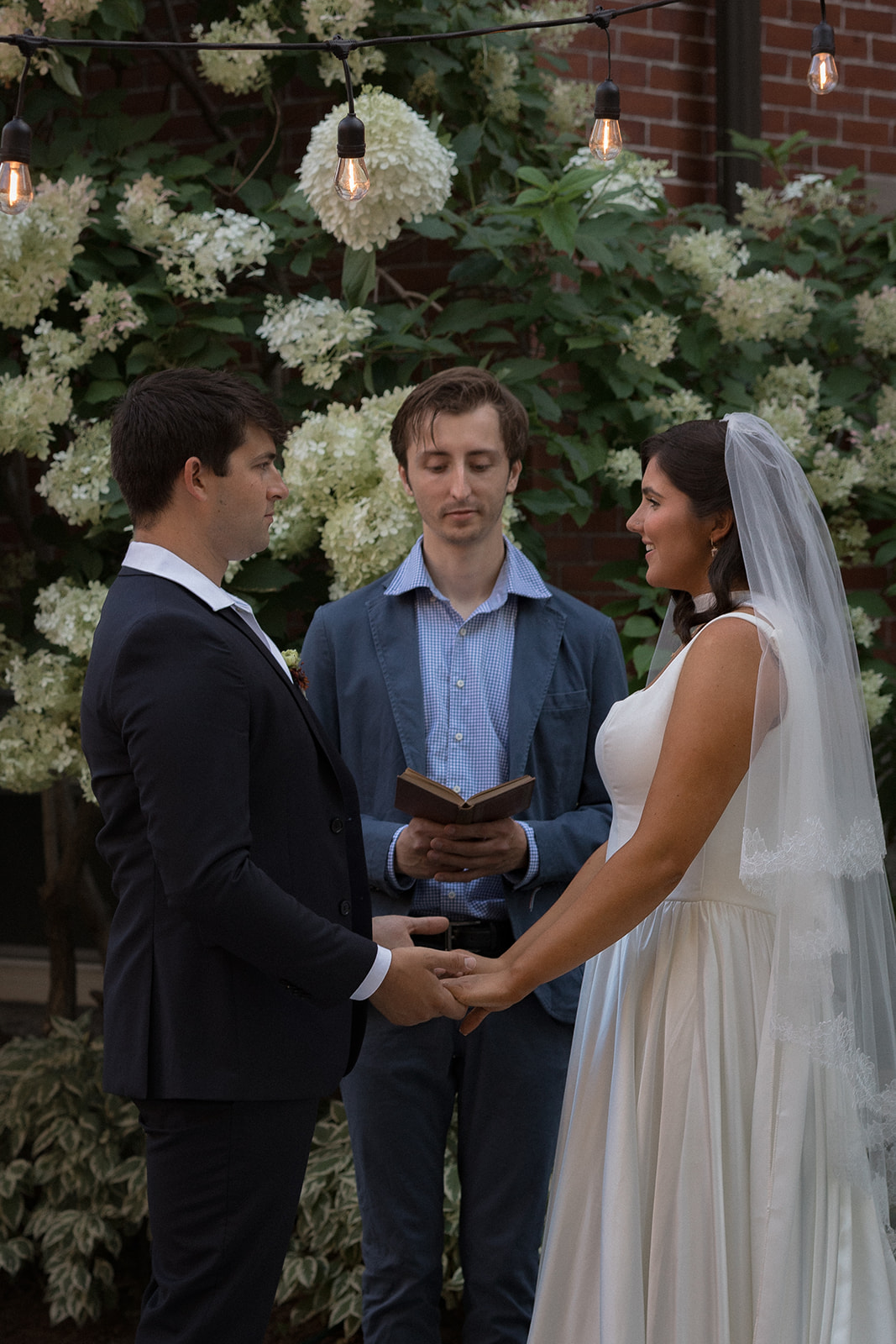 The couple holds hands while the officiant reads their vows beneath hanging café lights.
