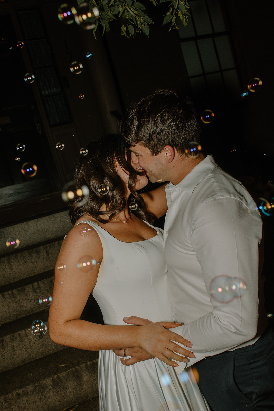 The couple sharing a kiss surrounded by floating bubbles under the porch light.
