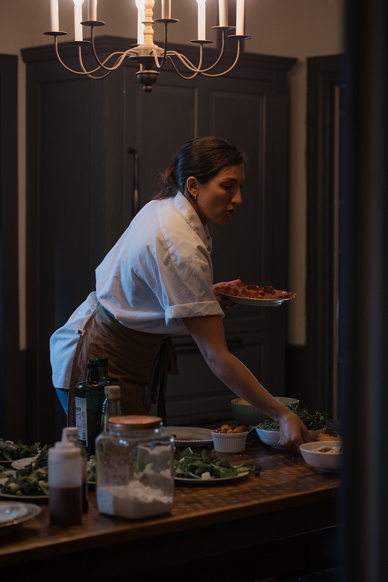 Chef preparing dinner, reaching across the table to finish plating salads beneath a glowing chandelier.