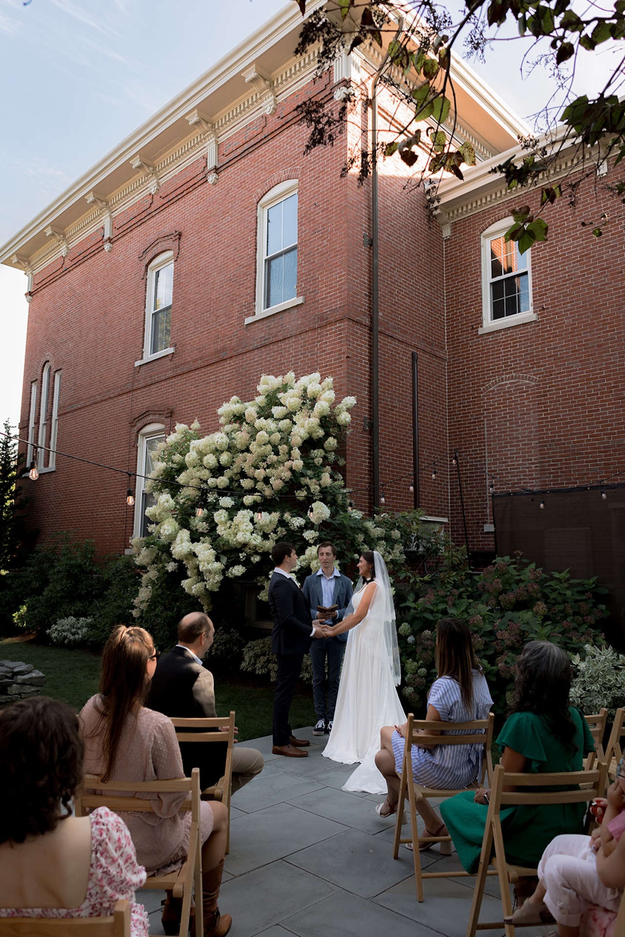 The couple exchanging vows outside a red brick building at one of Maine’s Historic Wedding Venues, guests seated around them under late-summer light.