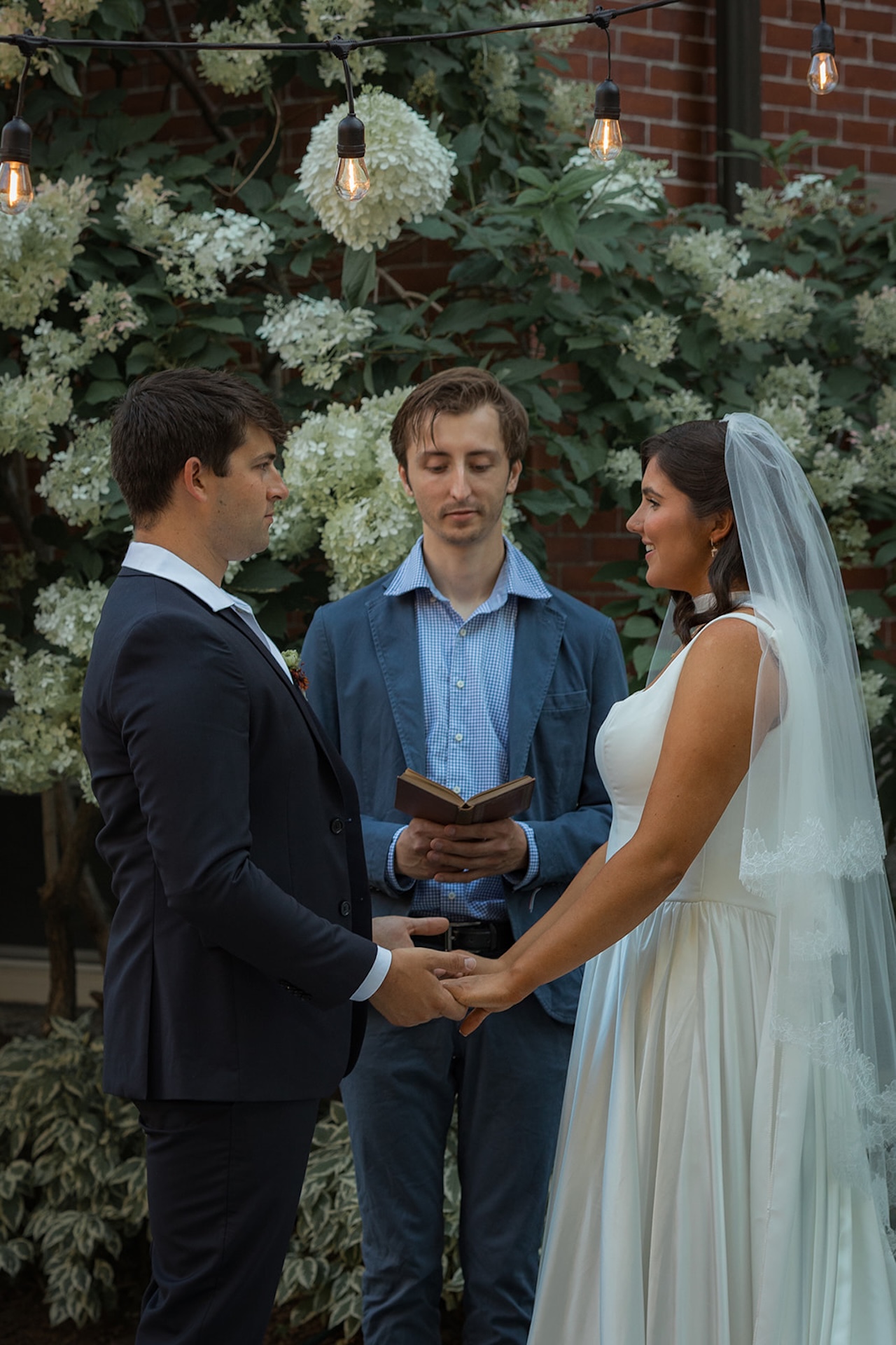 The couple holds hands while the officiant reads their vows beneath hanging café lights.
