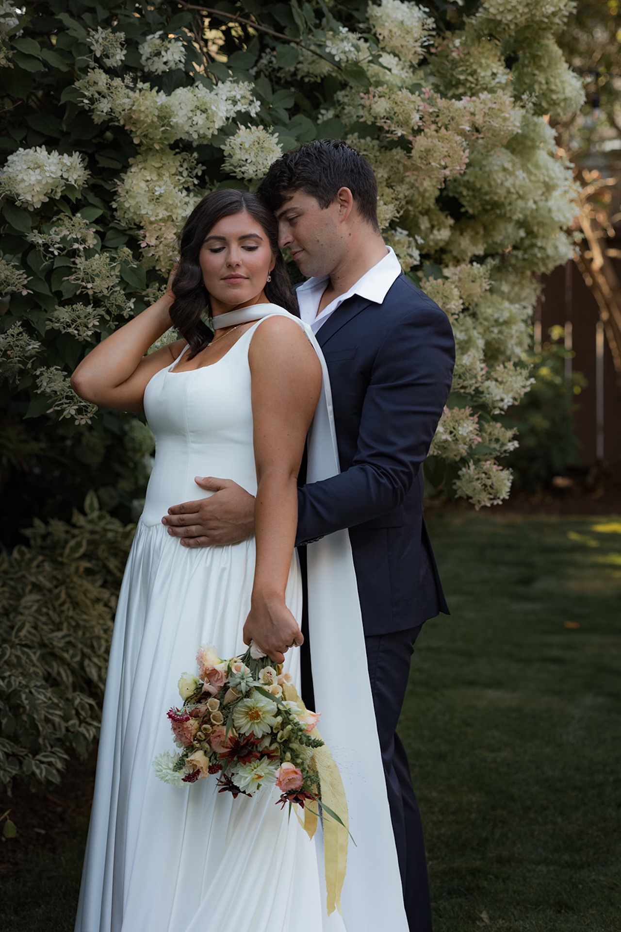 Bride and groom standing together in the garden, surrounded by soft white hydrangeas as they hold each other close.