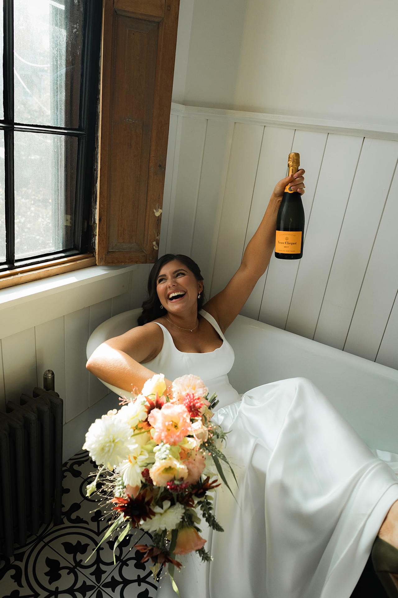 Bride laughing in a vintage clawfoot tub, holding up a bottle of Veuve Clicquot while her bouquet rests across her lap.