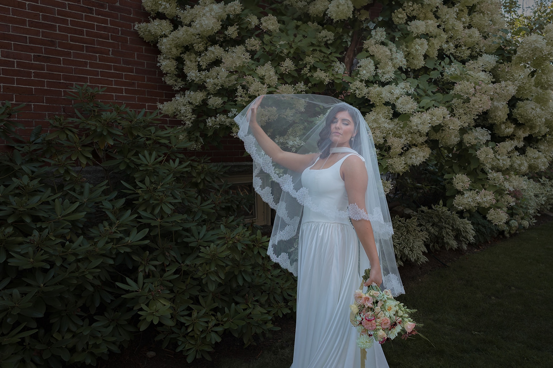 Bride standing in the garden holding her veil above her head, framed by blooming hydrangeas and soft evening light.