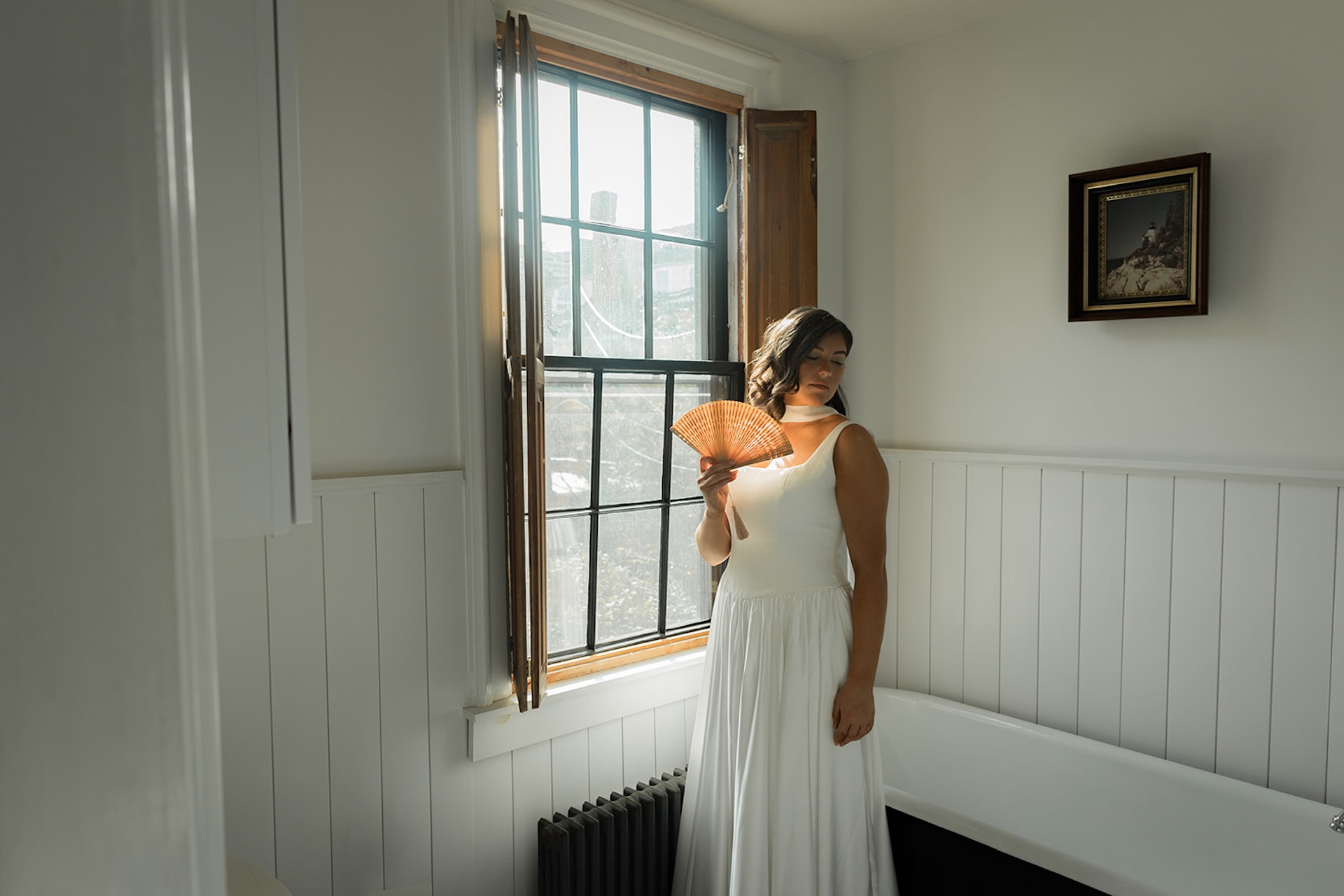 Bride standing by a tall window with her hand fan, sunlight streaming in and catching the gentle folds of her dress.