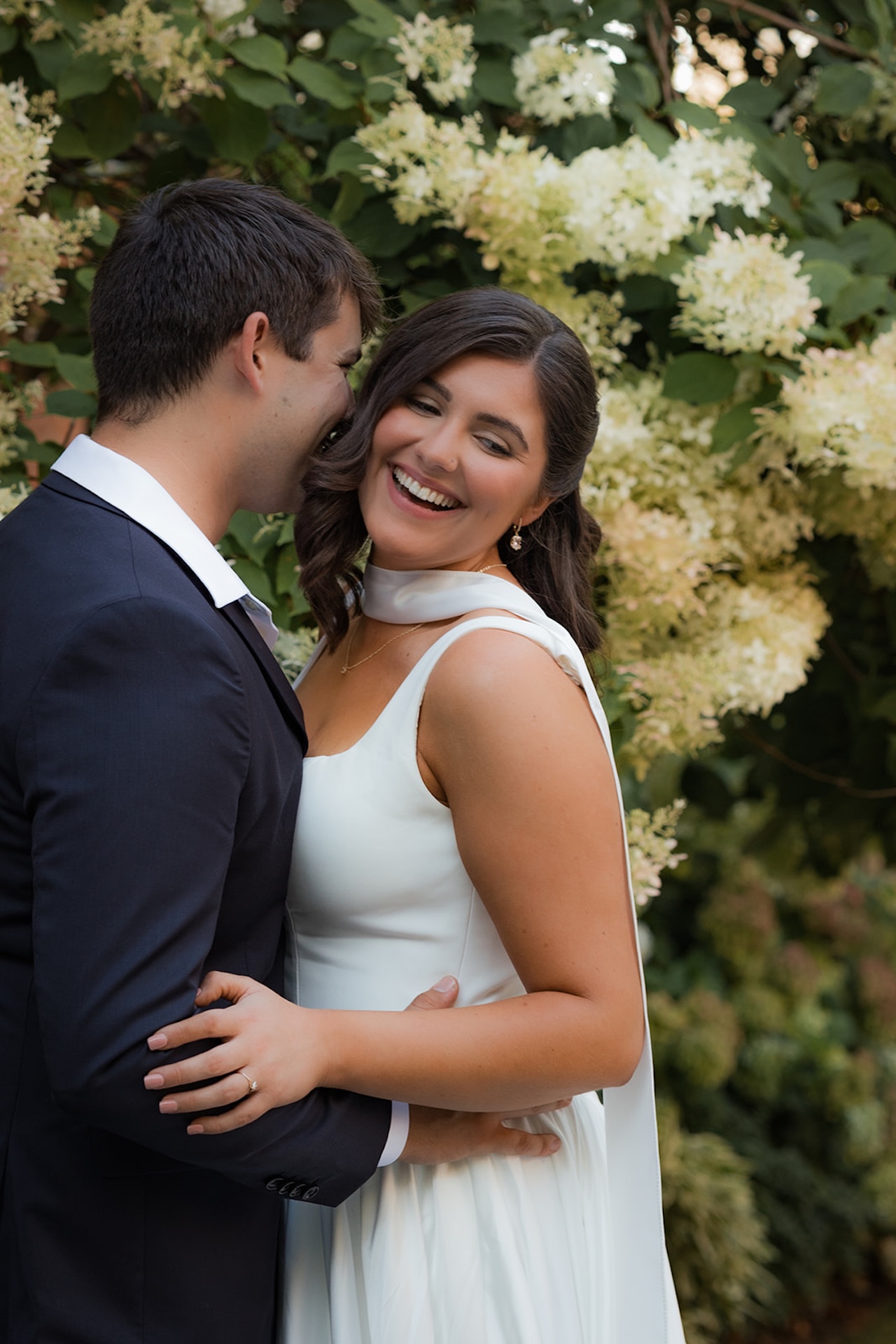 Groom kissing the bride’s cheek as she laughs, surrounded by lush hydrangeas on the garden lawn.