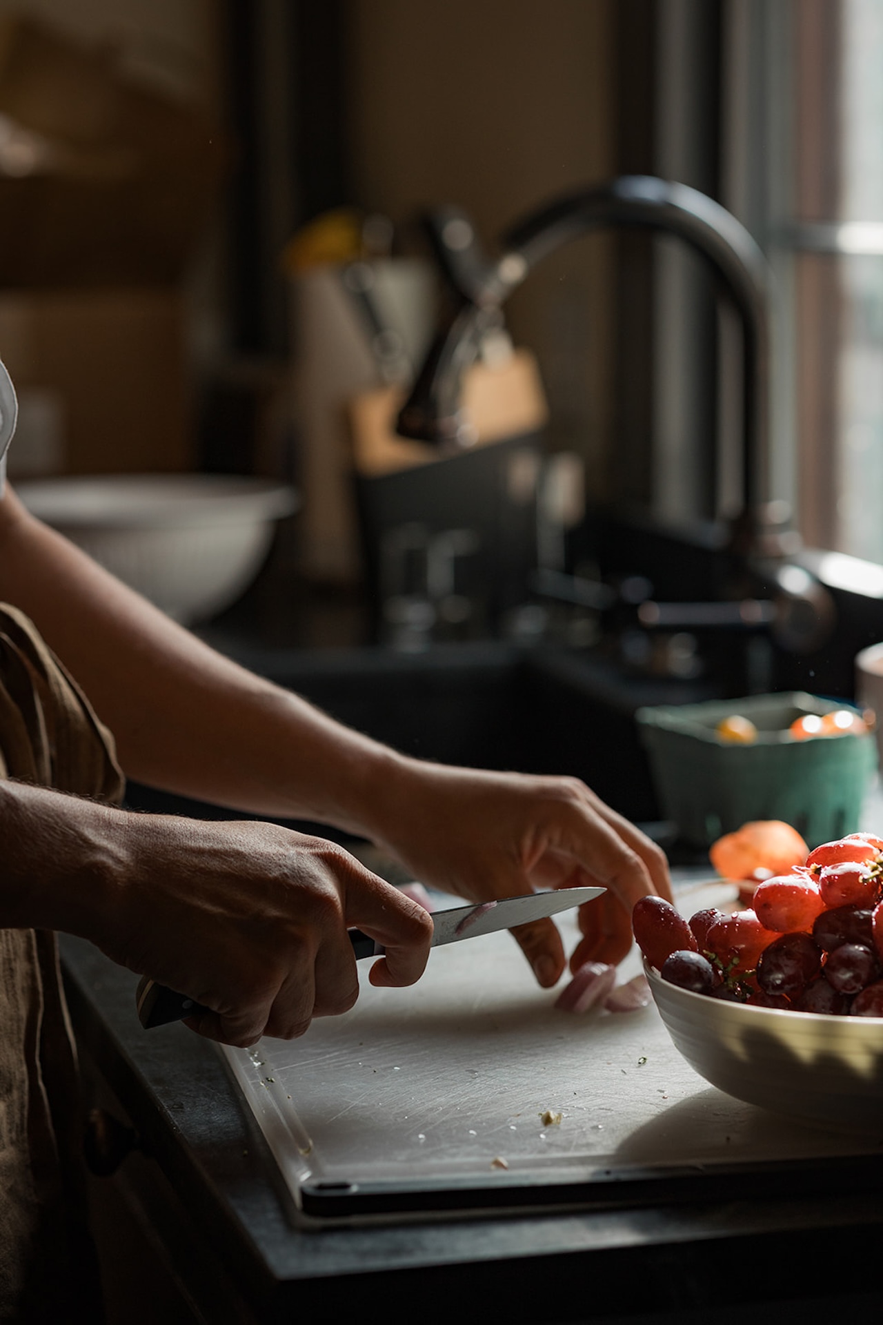 Private chef slicing red onions on a cutting board beside a bowl of grapes in warm kitchen light.