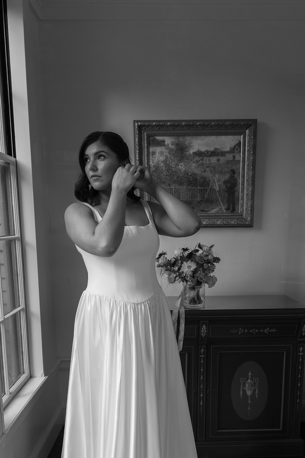 Bride putting on her earrings by the window, bouquet resting on the cabinet behind her.