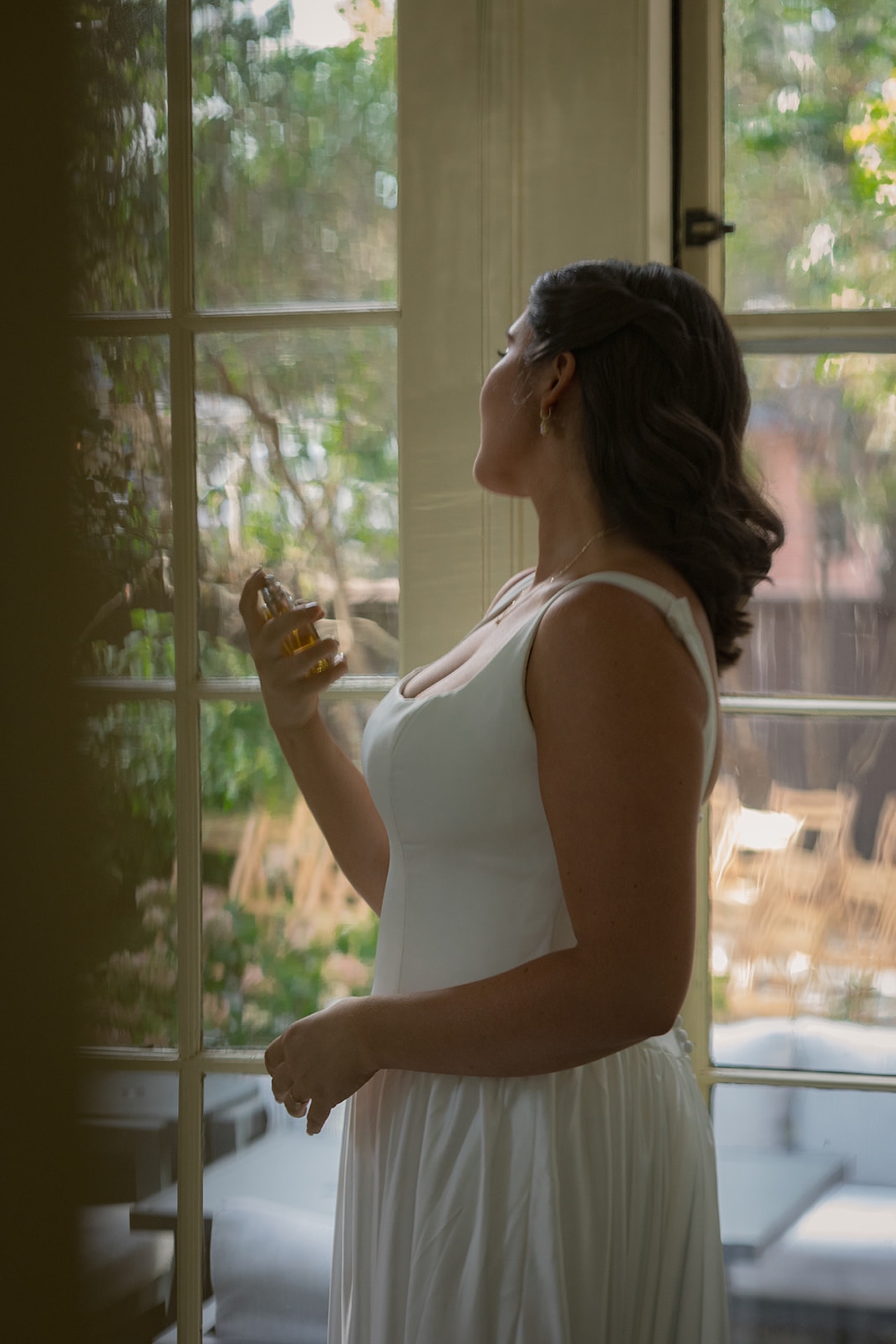 Bride spritzing perfume near a window, sunlight reflecting softly off her simple, elegant wedding dress.
