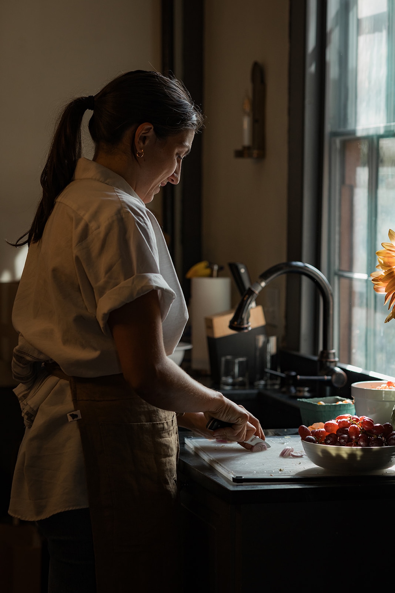 Chef smiling softly as she chops onions by the window, warm afternoon light falling across the counter.