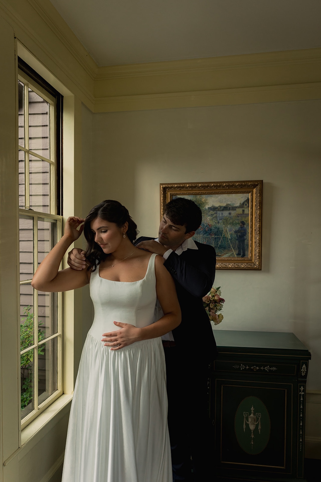 Groom fastening the bride’s necklace as she stands by a window, soft indoor light illuminating her dress inside one of Maine’s Historic Wedding Venues.